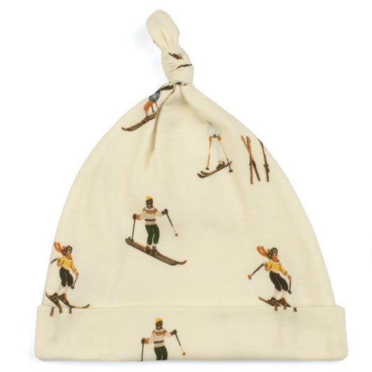 A beige organic cotton knotted baby hat with a vintage ski print, including illustrations of skiers and ski equipment.