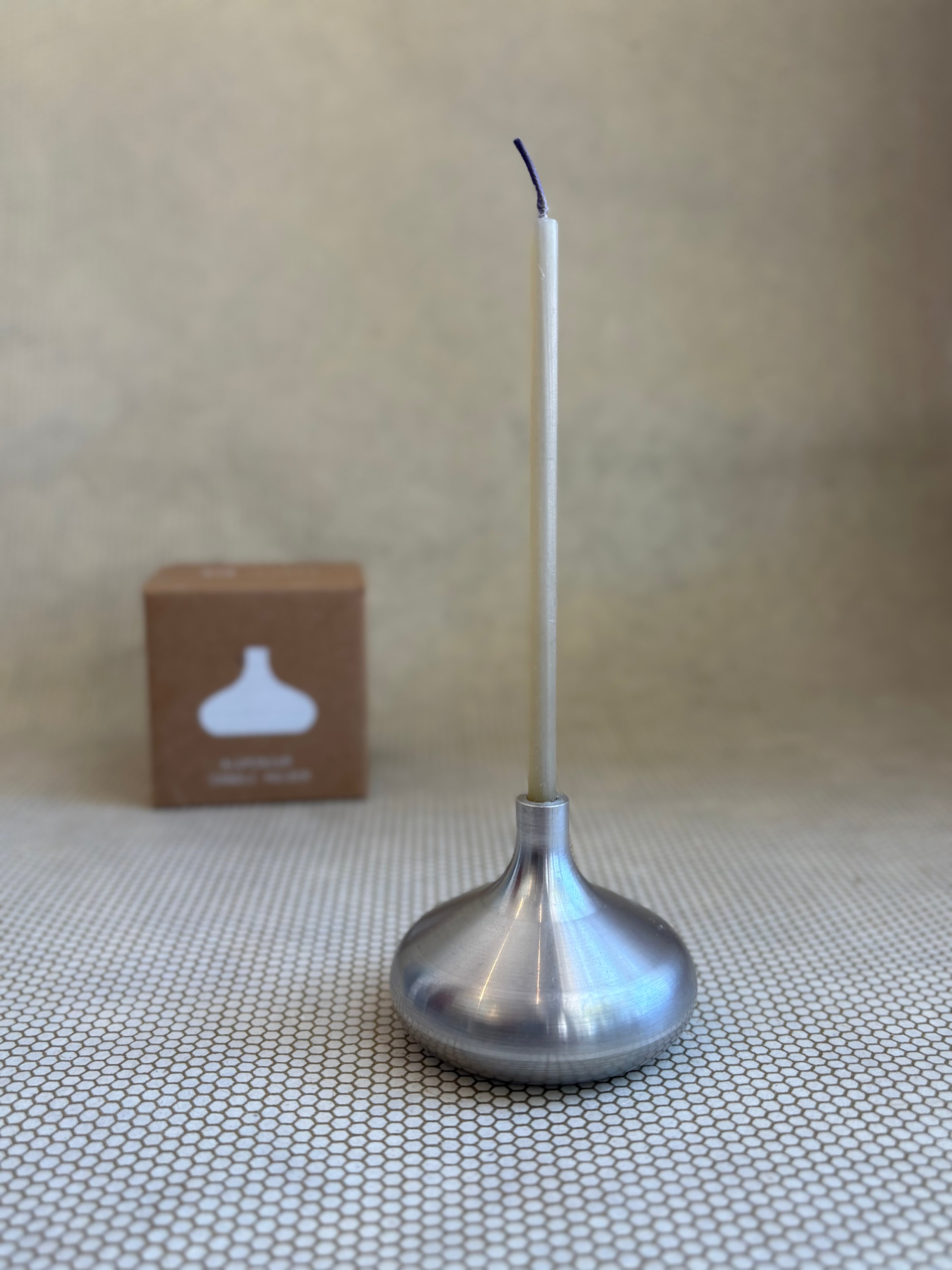 Silver candle holder with a white candle on a textured surface, with a brown box in the background.