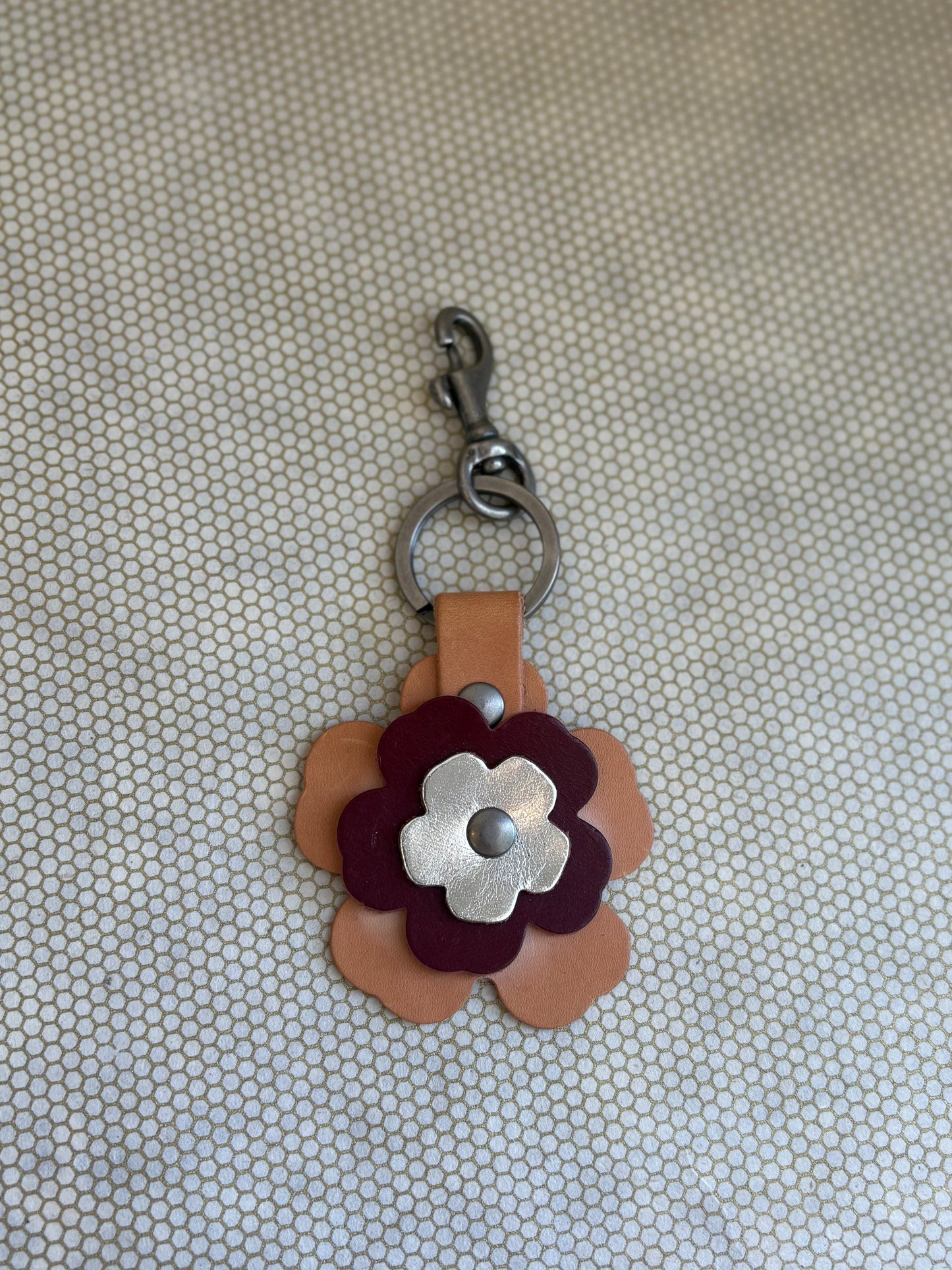 Floral-shaped keychain with a metal clasp on a textured beige background. metallic, wine, tan. 