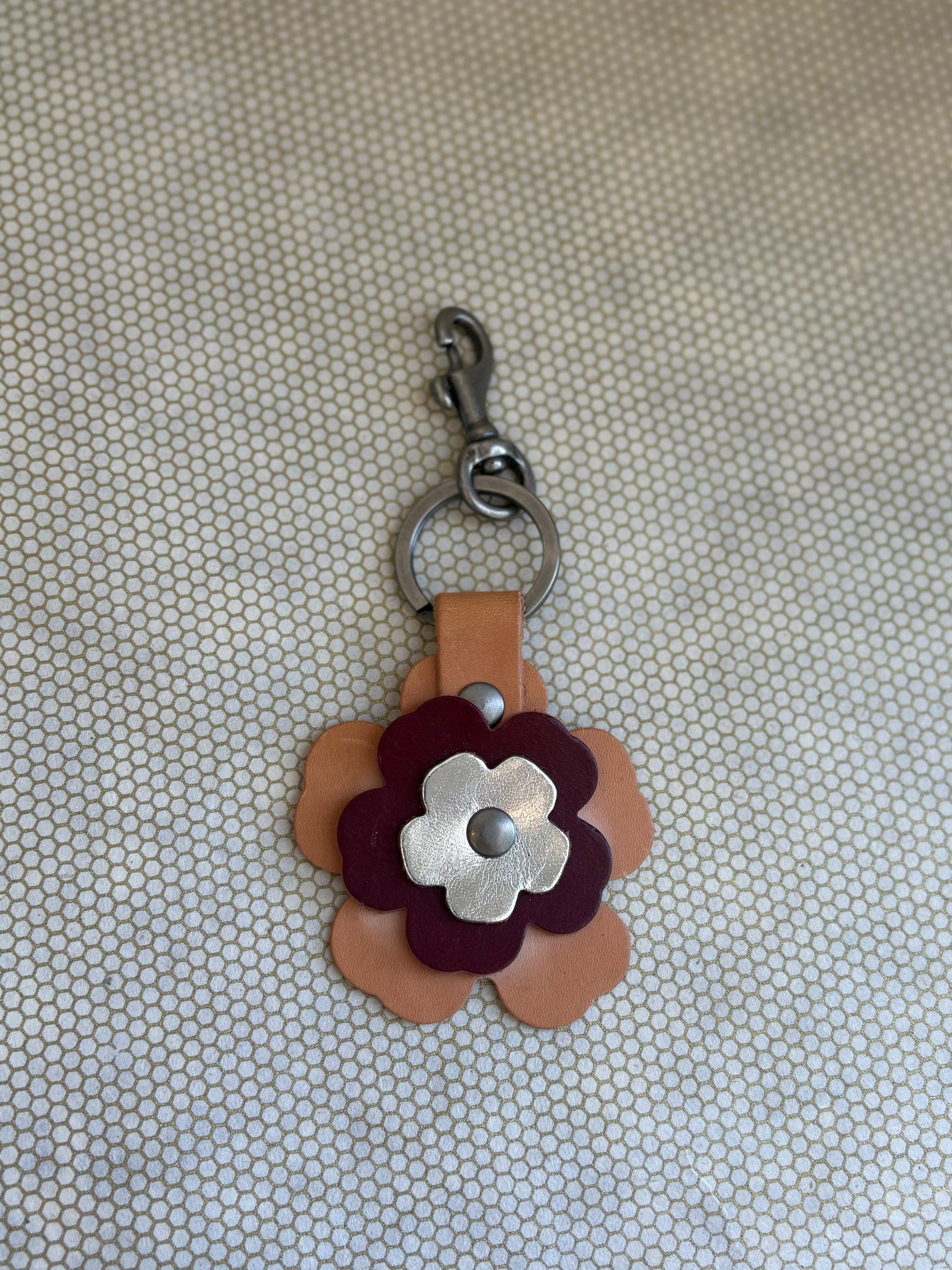 Floral-shaped keychain with a metal clasp on a textured beige background. metallic, wine, tan. 