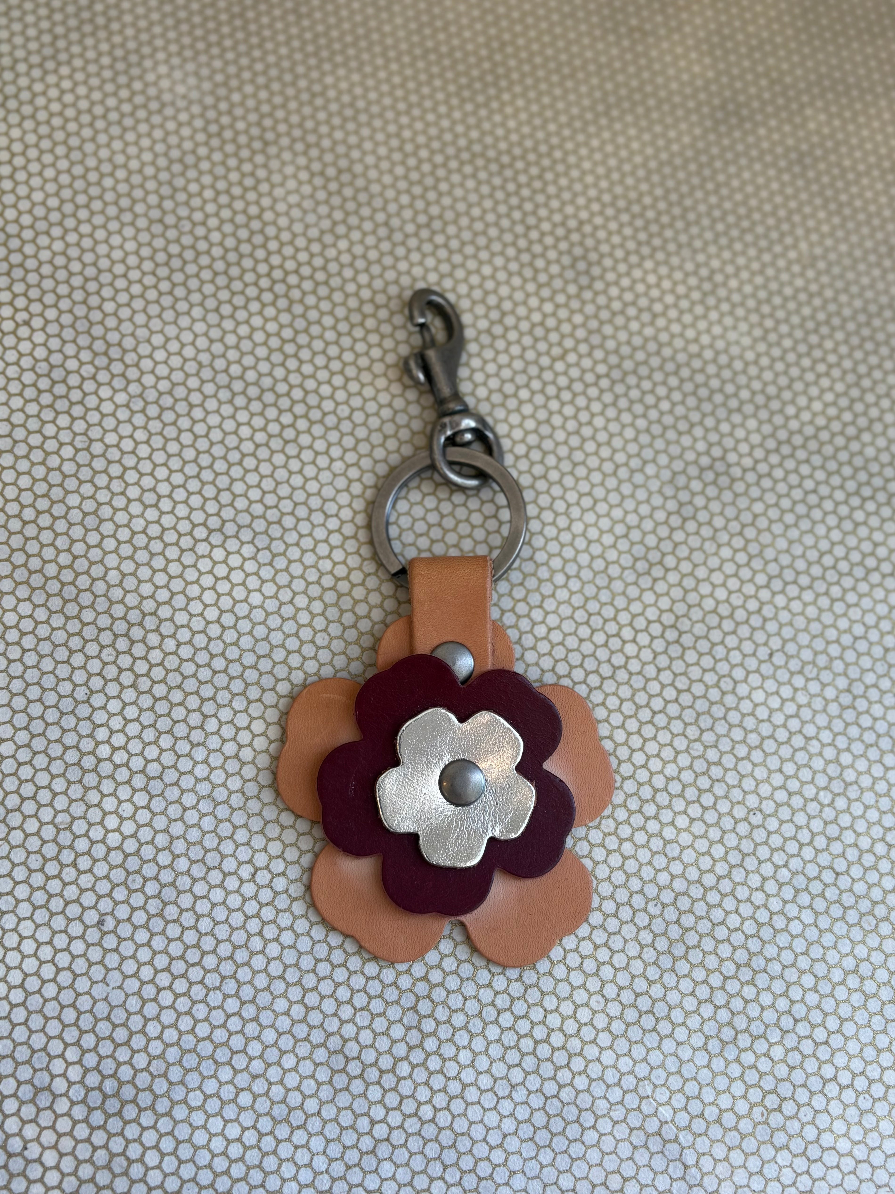 Floral-shaped keychain with a metal clasp on a textured beige background. metallic, wine, tan. 