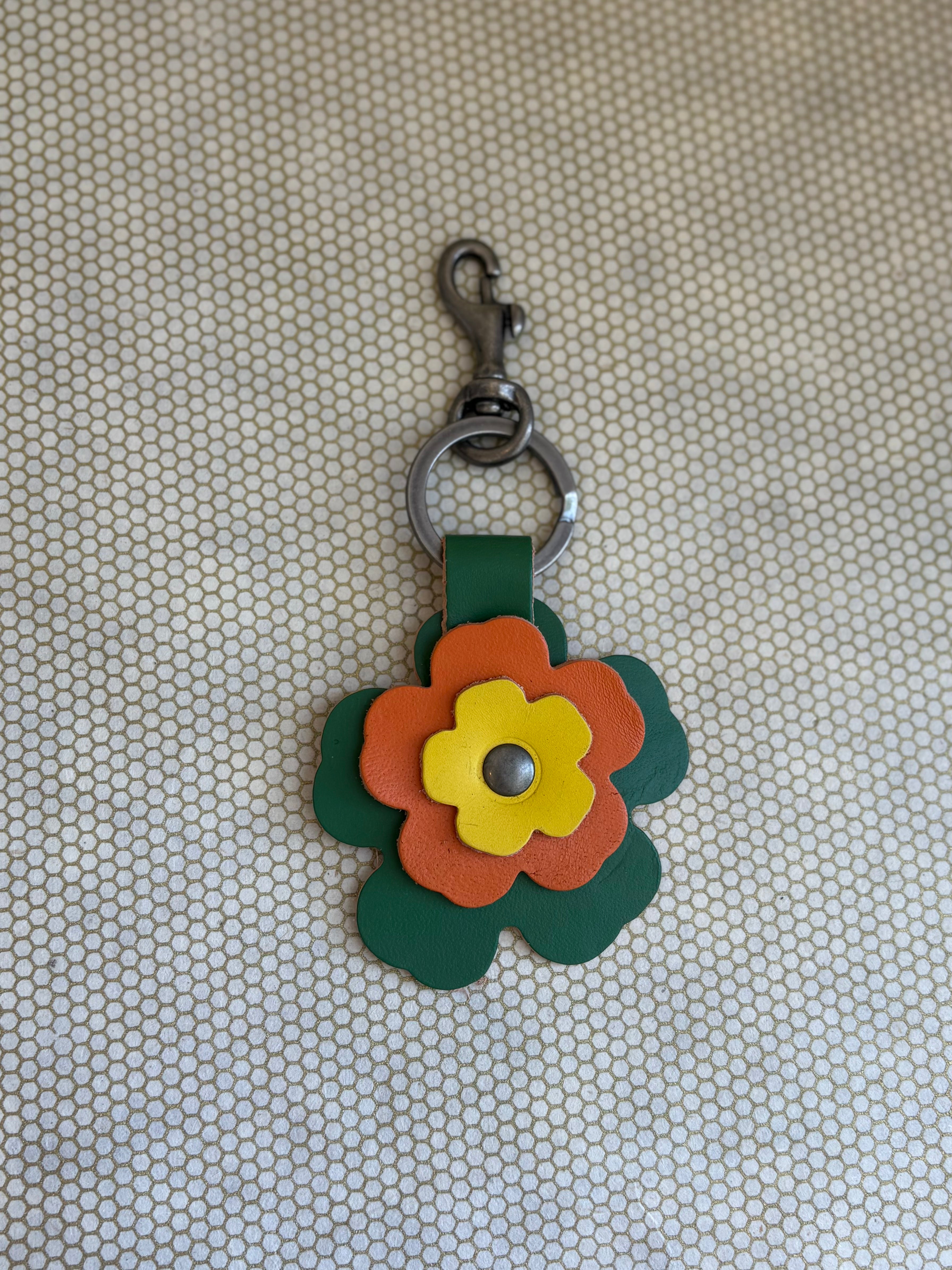Floral-shaped keychain with a metal clasp on a textured beige background. yellow, orange, green. 