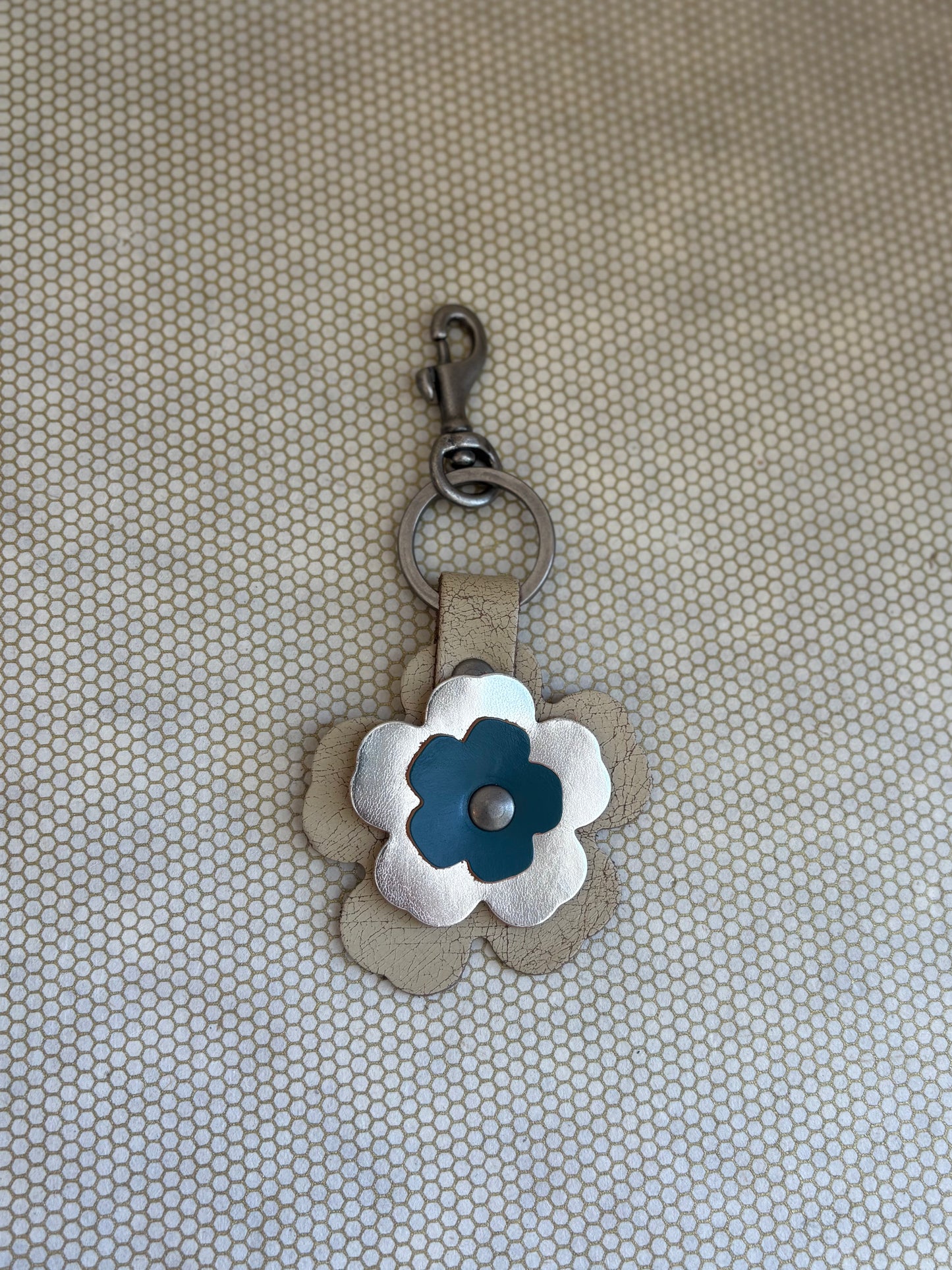 Floral-shaped keychain with a metal clasp on a textured beige background. metallic, blue, neutral. 