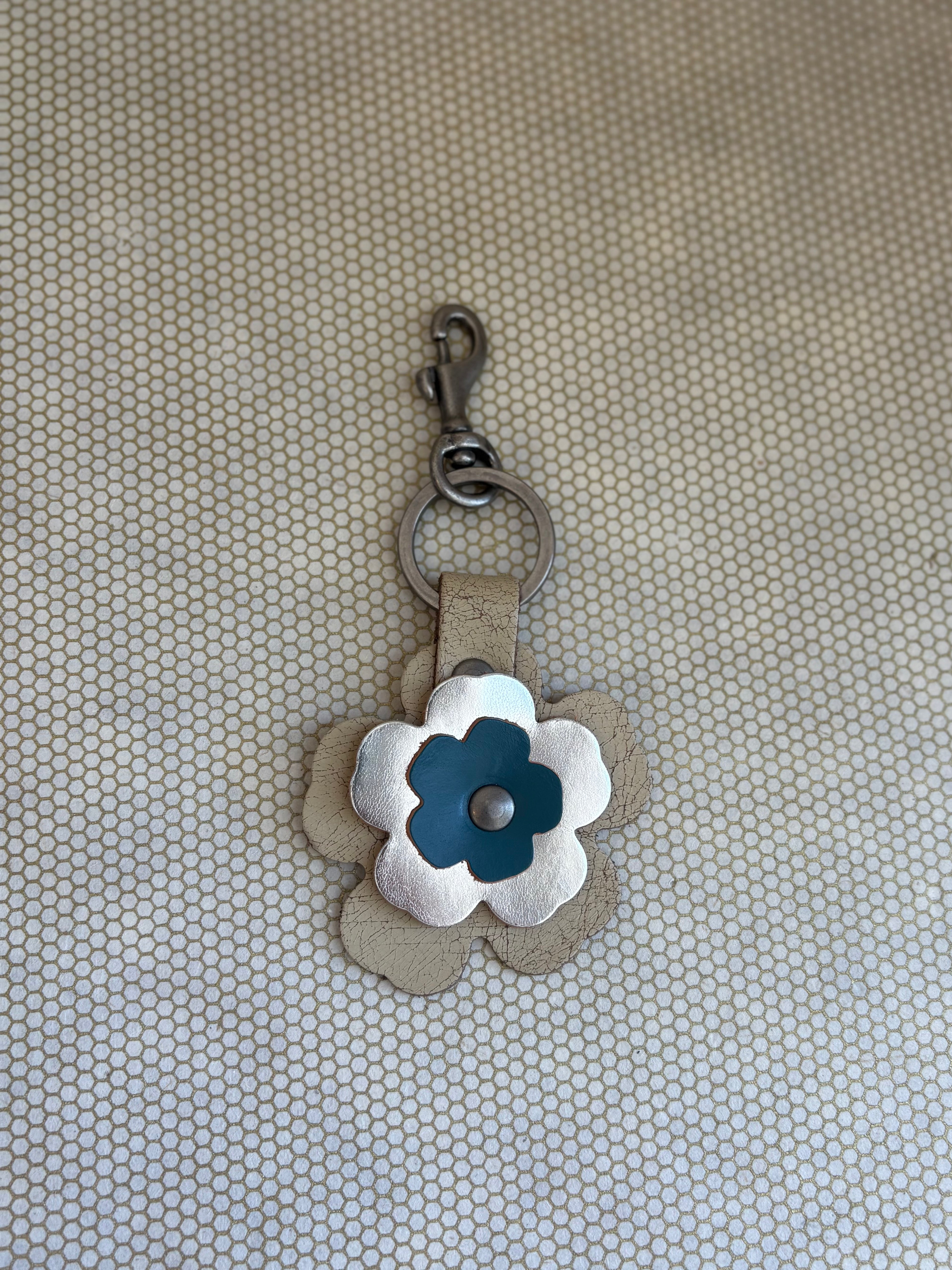 Floral-shaped keychain with a metal clasp on a textured beige background. metallic, blue, neutral. 