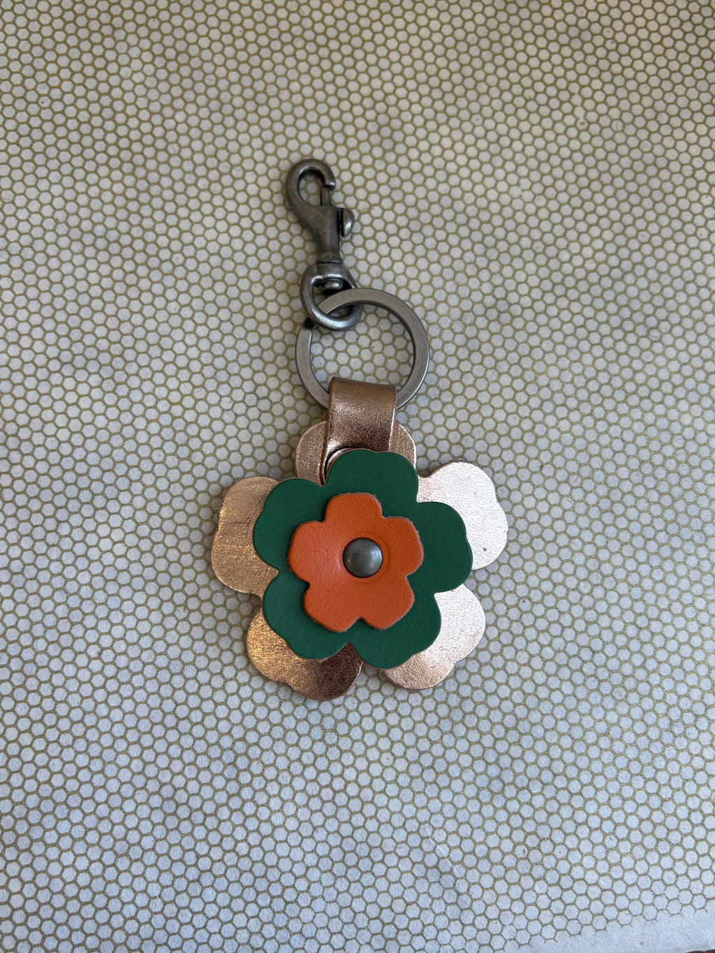 Floral-shaped keychain with a metal clasp on a textured beige background. metallic, orange, green. 
