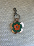Floral-shaped keychain with a metal clasp on a textured beige background. metallic, orange, green. 