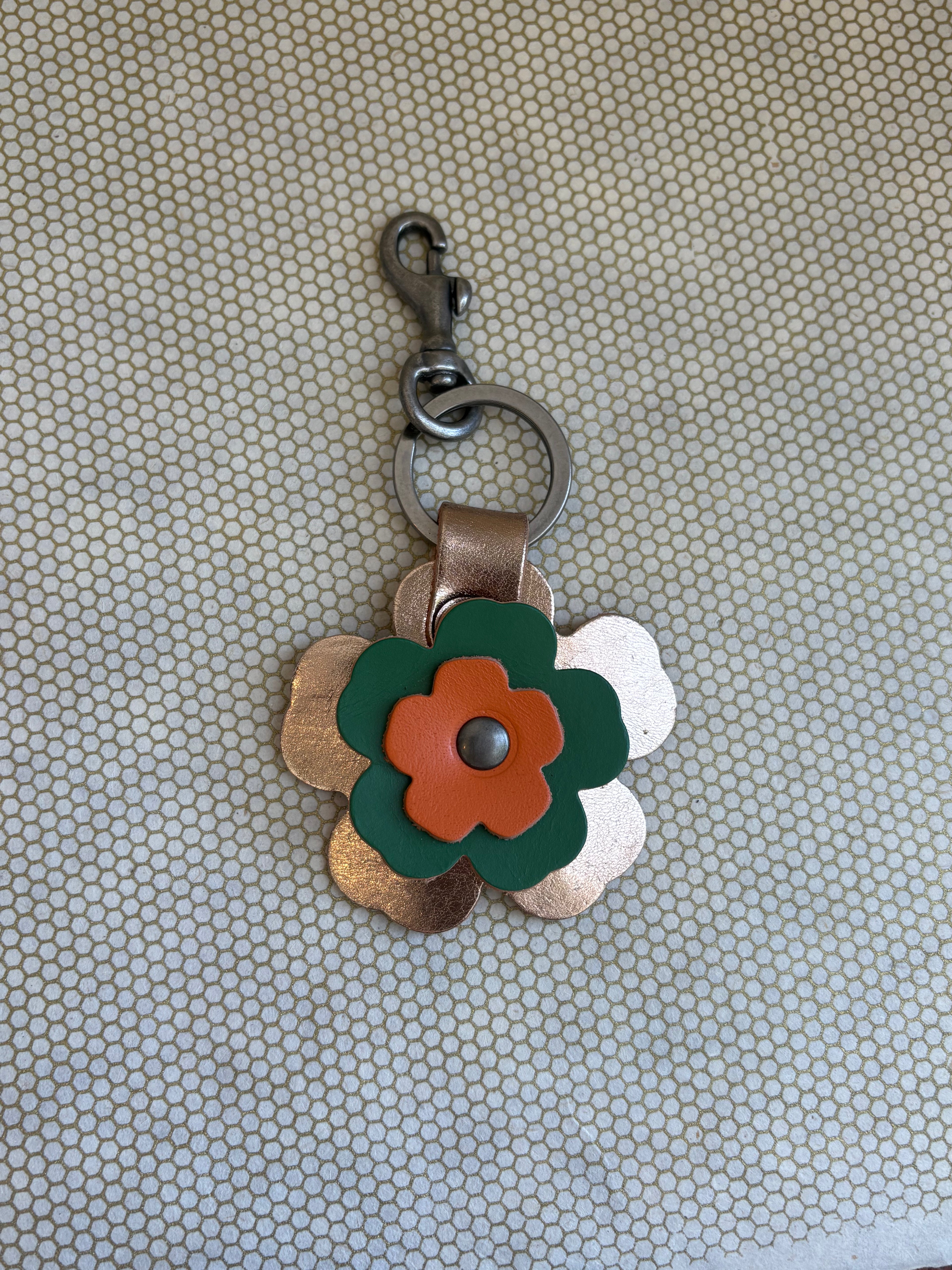 Floral-shaped keychain with a metal clasp on a textured beige background. metallic, orange, green. 