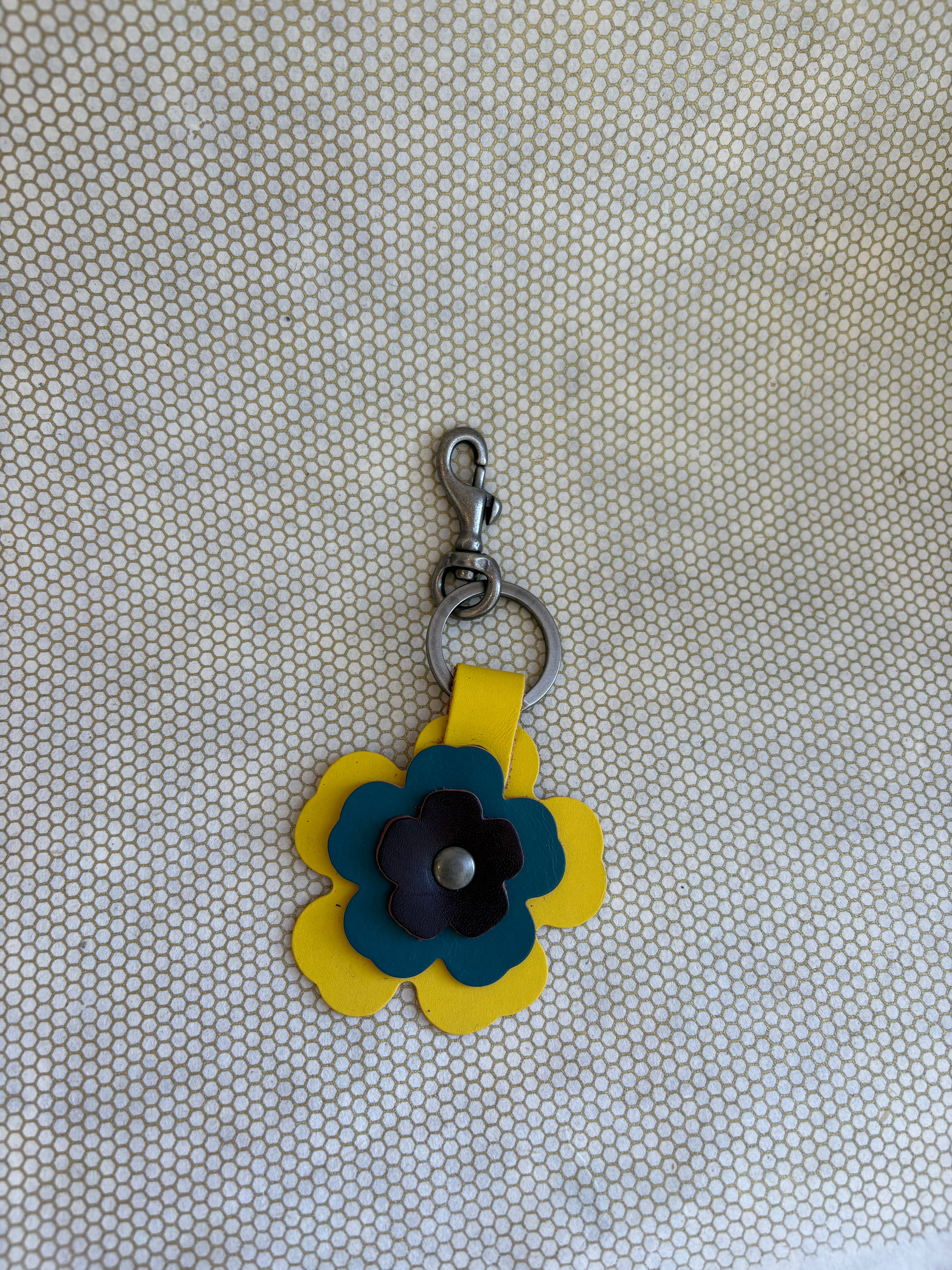 Floral-shaped keychain with a metal clasp on a textured beige background. blue, yellow, chocolate. 
