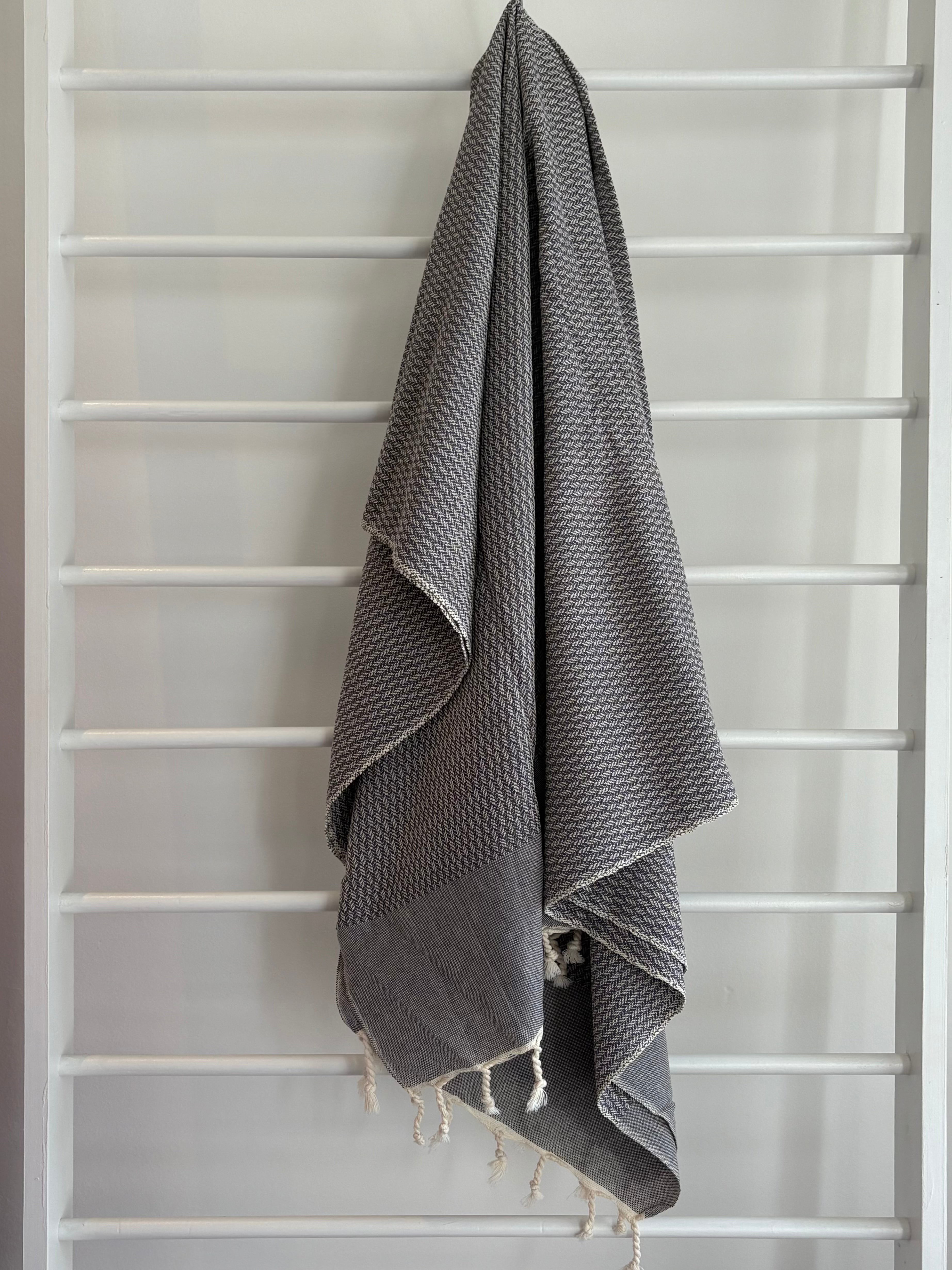 Gray textured towel with tassels hanging on a white ladder. dark grey tweed weave towel. 