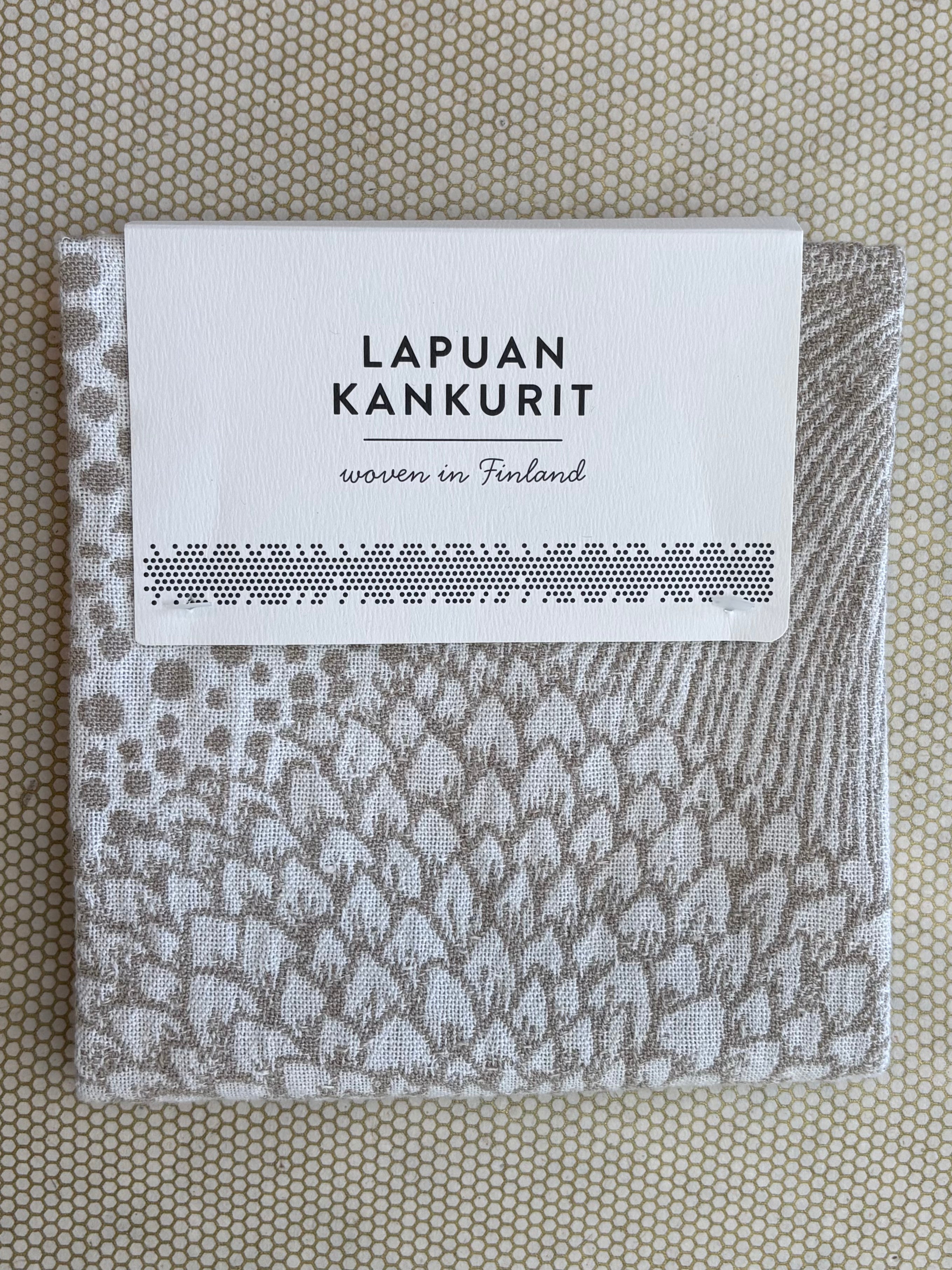 A folded woven textile with a gray and off-white leaf pattern, wrapped in a white paper label that reads 'LAPUAN KANKURIT' and 'woven in Finland', placed on a beige honeycomb-patterned surface.