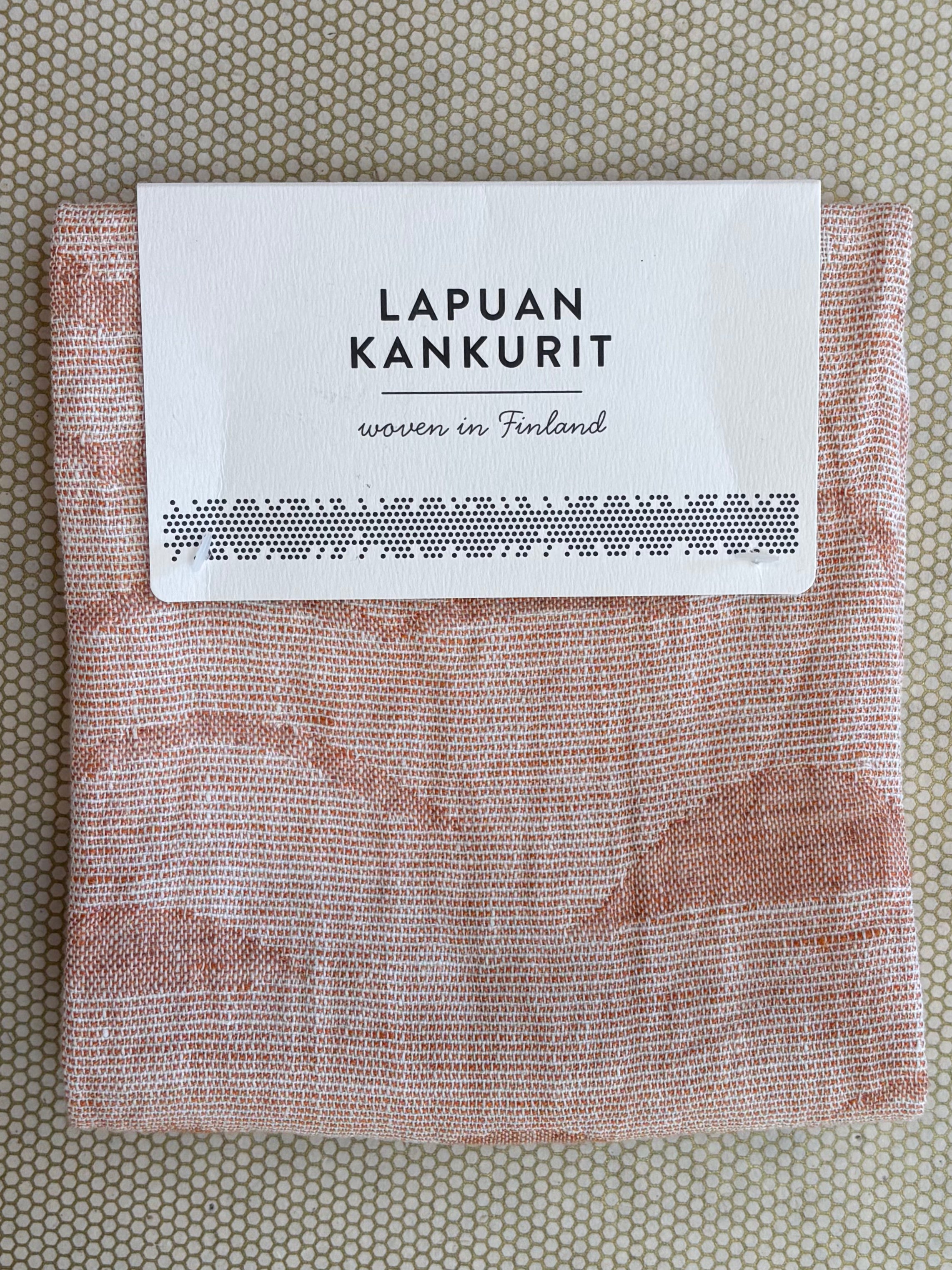 A folded white paper label reading 'LAPUAN KANKURIT' and 'woven in Finland' placed on top of an orange and white patterned kitchen towel, photographed from above on a beige textured surface.