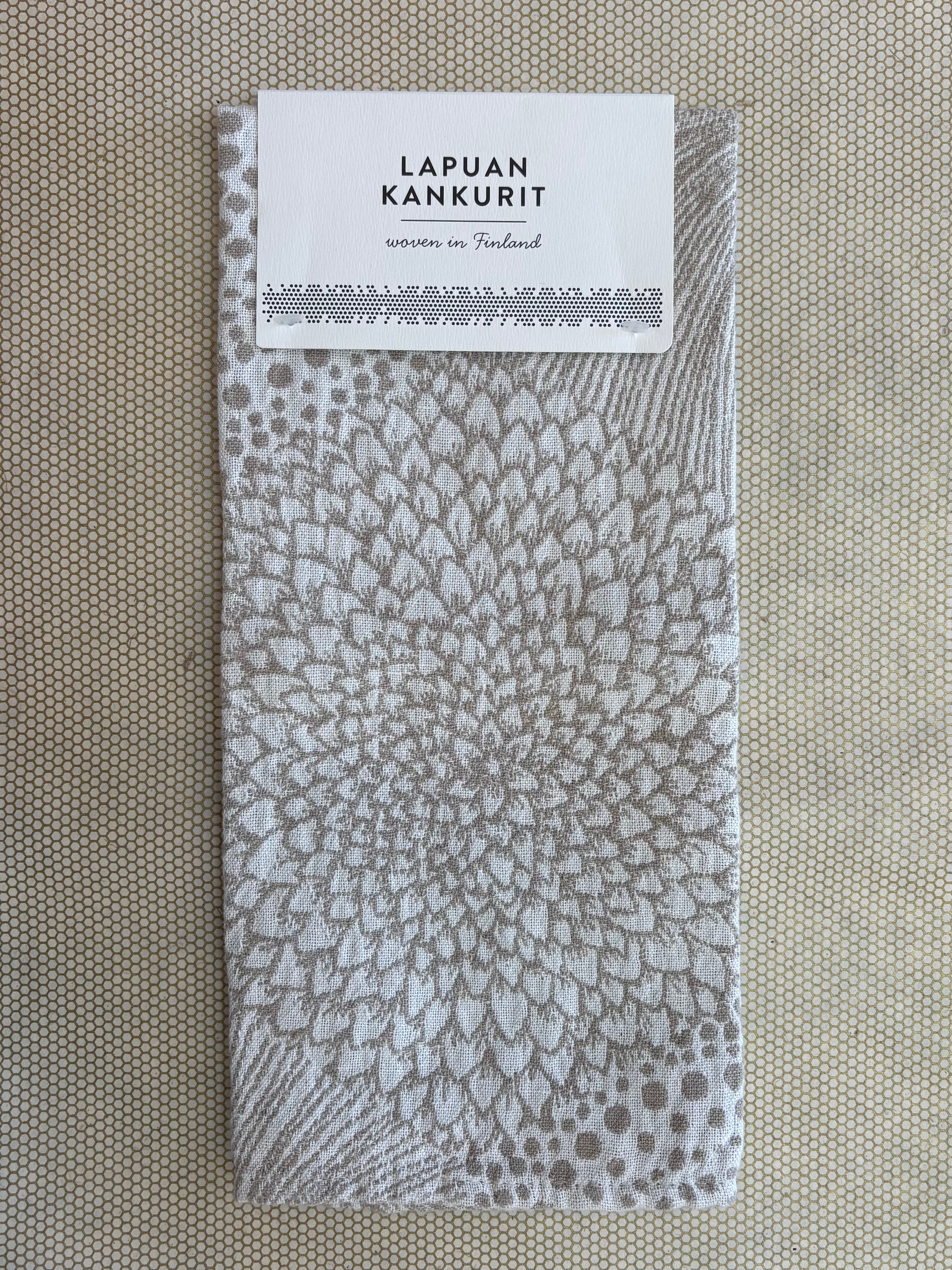 A square woven fabric napkin with a gray and white radial floral pattern, featuring a white paper label at the top that reads 'LAPUAN KANKURIT' and 'woven in Finland'. The napkin is laid flat on a textured beige surface.