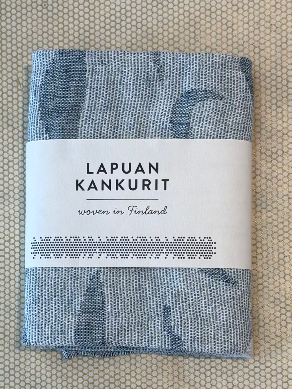 A folded light blue and white patterned woven hand or kitchen towel, wrapped with a white paper label that reads 'LAPUAN KANKURIT' and 'woven in Finland,' placed on a beige patterned surface.