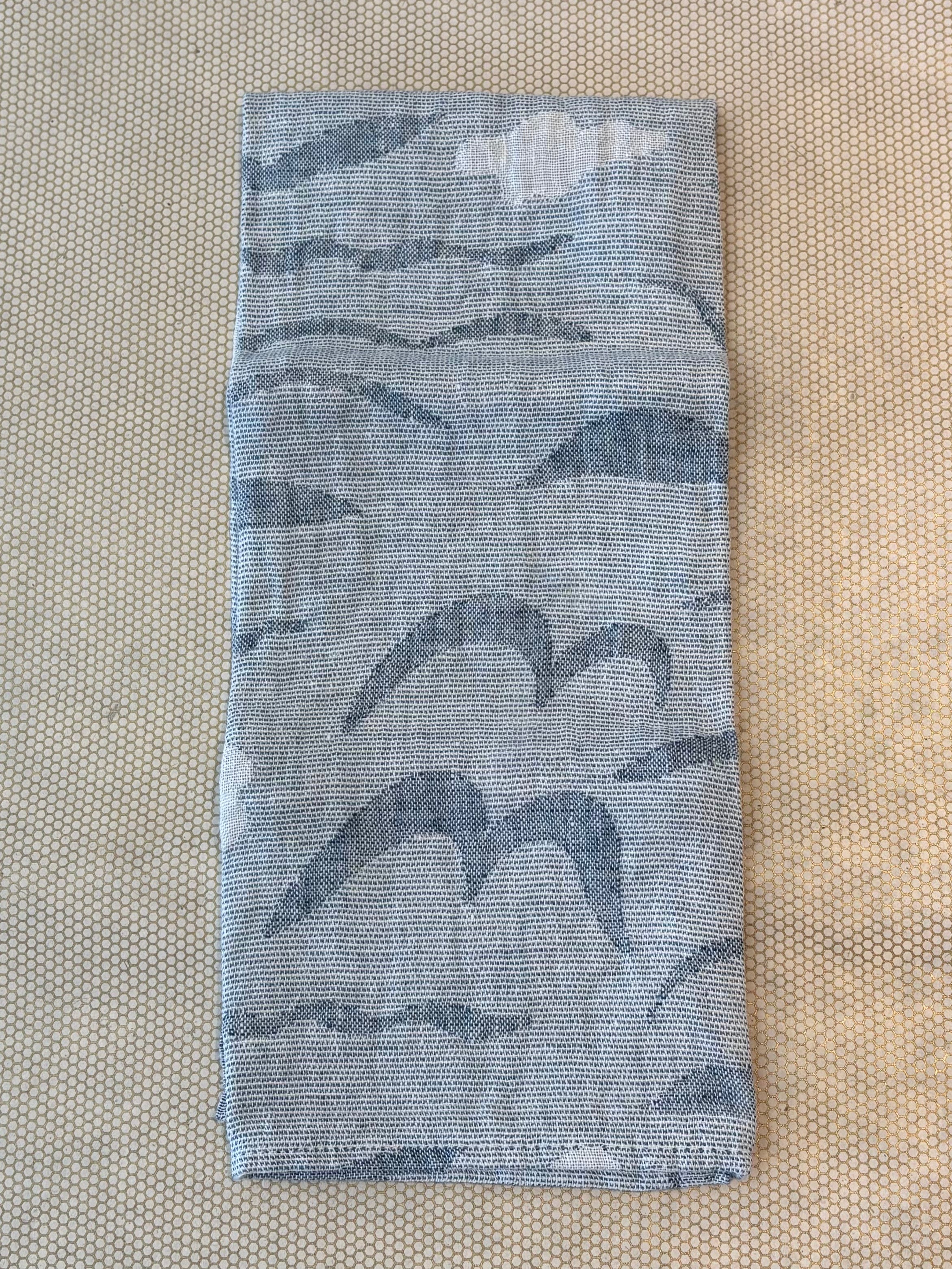 A folded light blue and white patterned woven hand or kitchen towel, wrapped with a white paper label that reads 'LAPUAN KANKURIT' and 'woven in Finland,' placed on a beige patterned surface.