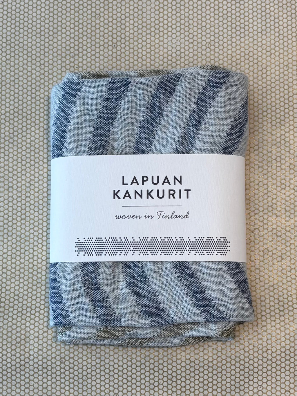 A folded woven textile with blue-gray and off-white striped pattern, wrapped in a white paper label that reads 'LAPUAN KANKURIT' and 'woven in Finland', placed on a textured beige surface.