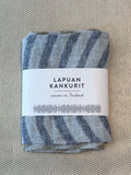 A folded woven textile with blue-gray and off-white striped pattern, wrapped in a white paper label that reads 'LAPUAN KANKURIT' and 'woven in Finland', placed on a textured beige surface.