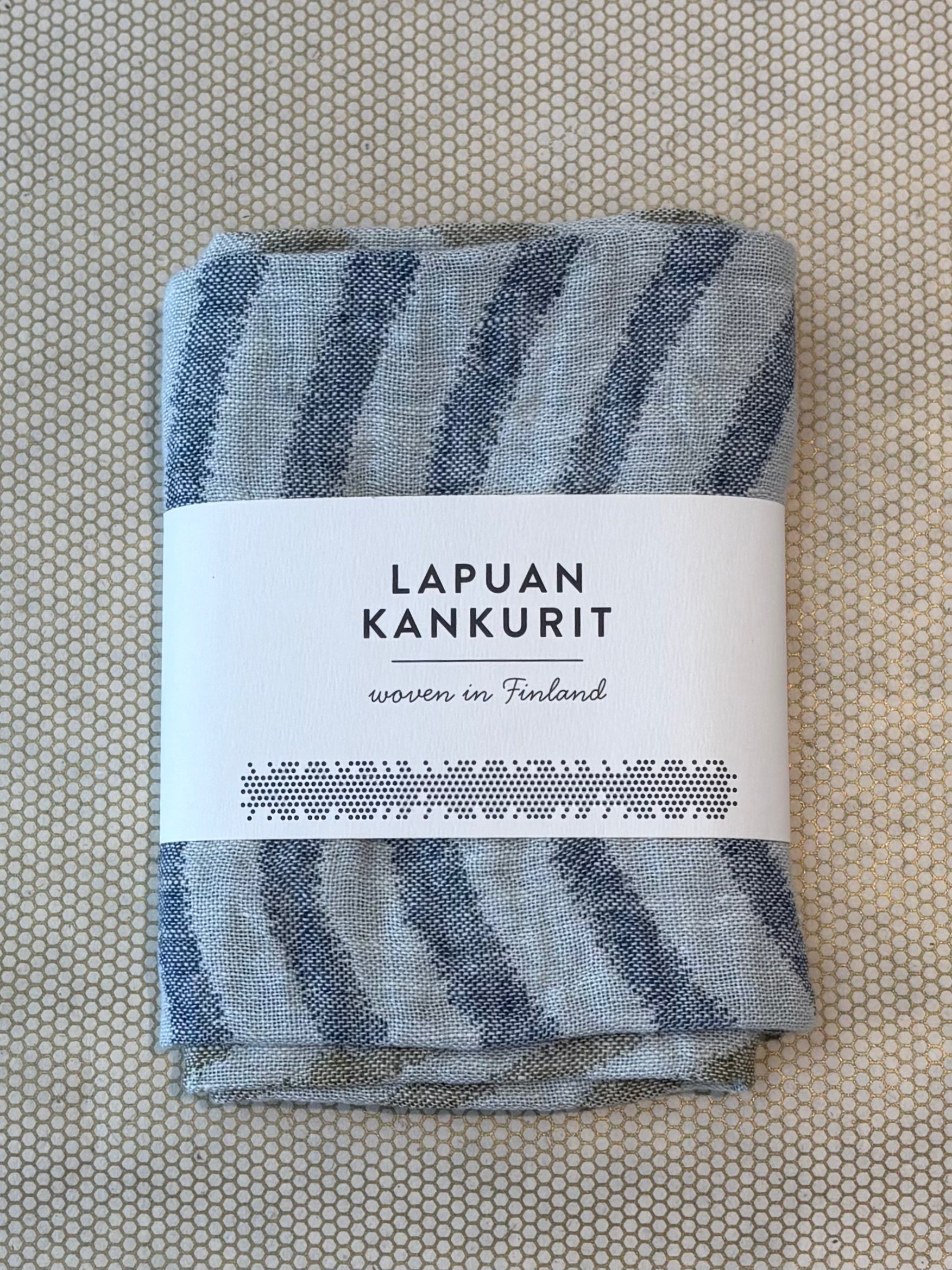 A folded woven textile with blue-gray and off-white striped pattern, wrapped in a white paper label that reads 'LAPUAN KANKURIT' and 'woven in Finland', placed on a textured beige surface.
