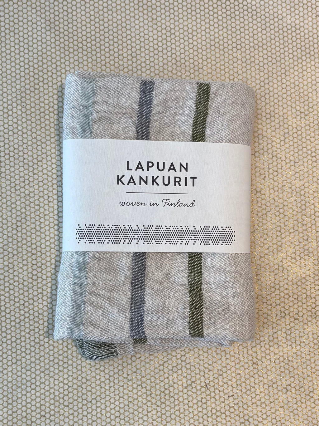 A folded light gray woven textile kitchen towel with dark gray vertical stripes, wrapped in a white paper label that reads 'LAPUAN KANKURIT' and 'woven in Finland,' placed on a textured beige surface.
