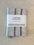 A folded light gray woven textile kitchen towel with dark gray vertical stripes, wrapped in a white paper label that reads 'LAPUAN KANKURIT' and 'woven in Finland,' placed on a textured beige surface.