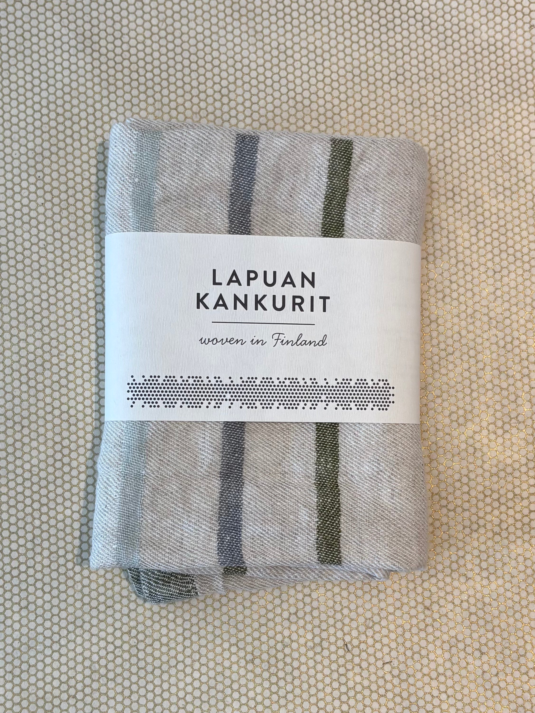 A folded light gray woven textile kitchen towel with dark gray vertical stripes, wrapped in a white paper label that reads 'LAPUAN KANKURIT' and 'woven in Finland,' placed on a textured beige surface.