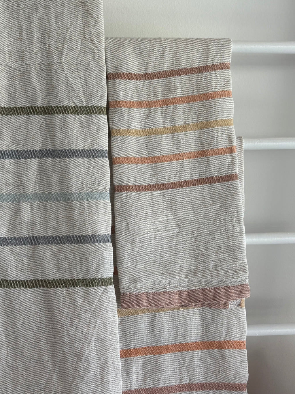 Two textured linen kitchen towels with horizontal stripes in muted earthy tones, hanging on a white towel rack.
