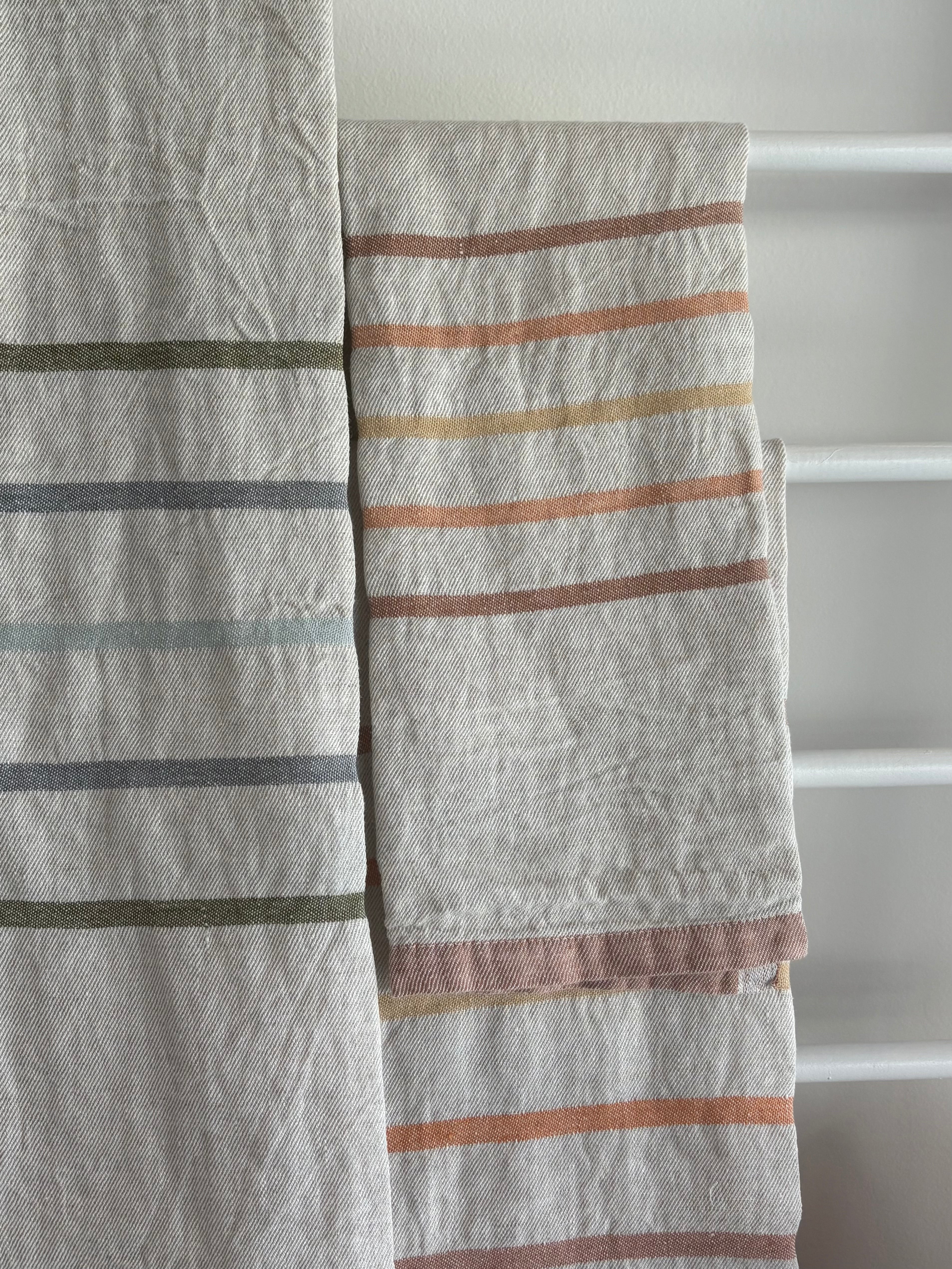 Two textured linen kitchen towels with horizontal stripes in muted earthy tones, hanging on a white towel rack.