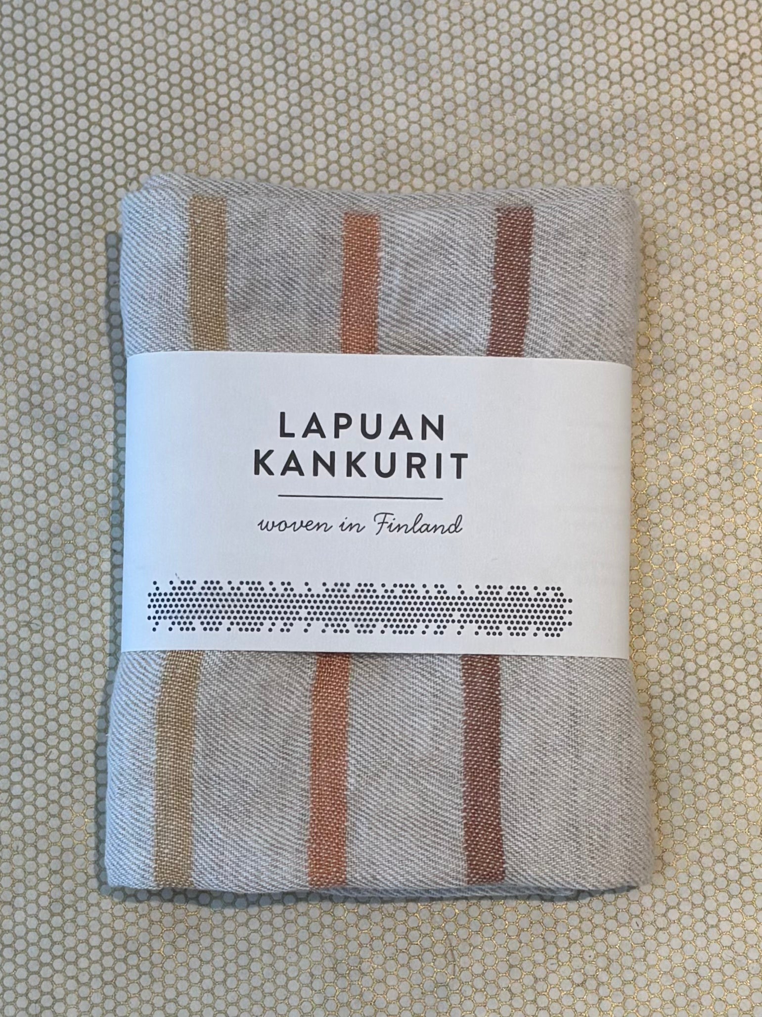 A folded woven Lapuan Kankurit kitchen towel with vertical stripes in beige, brown, and muted orange, wrapped in a white paper label that reads 'LAPUAN KANKURIT' and 'woven in Finland'.
