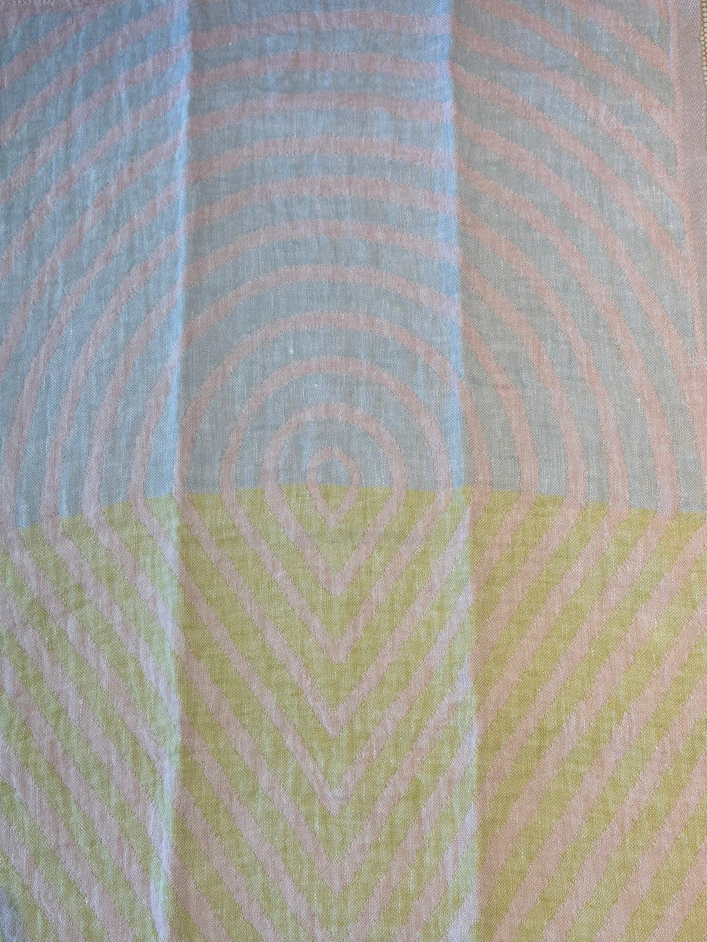 A close-up of a textured fabric with a geometric pattern, featuring concentric circles in light blue and pale pink on the top half, and chevron lines in pale yellow and pink on the bottom half.