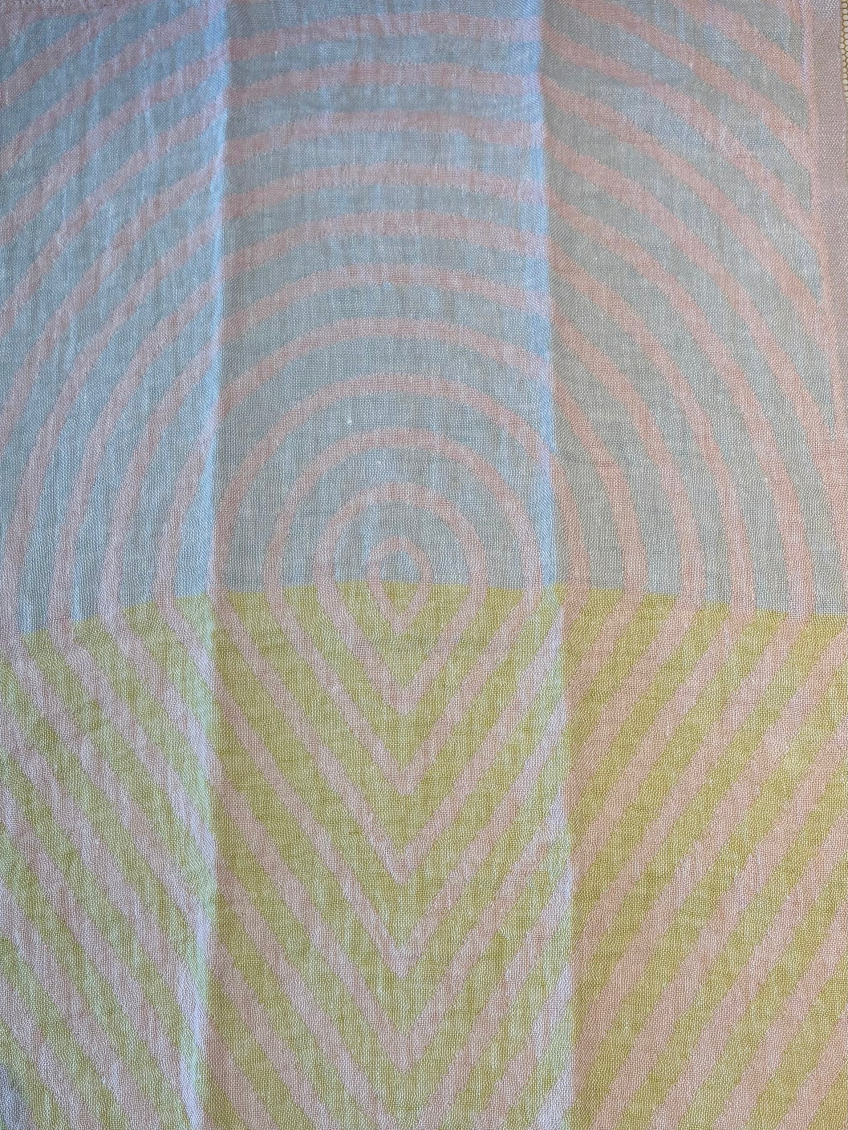 A close-up of a textured fabric with a geometric pattern, featuring concentric circles in light blue and pale pink on the top half, and chevron lines in pale yellow and pink on the bottom half.