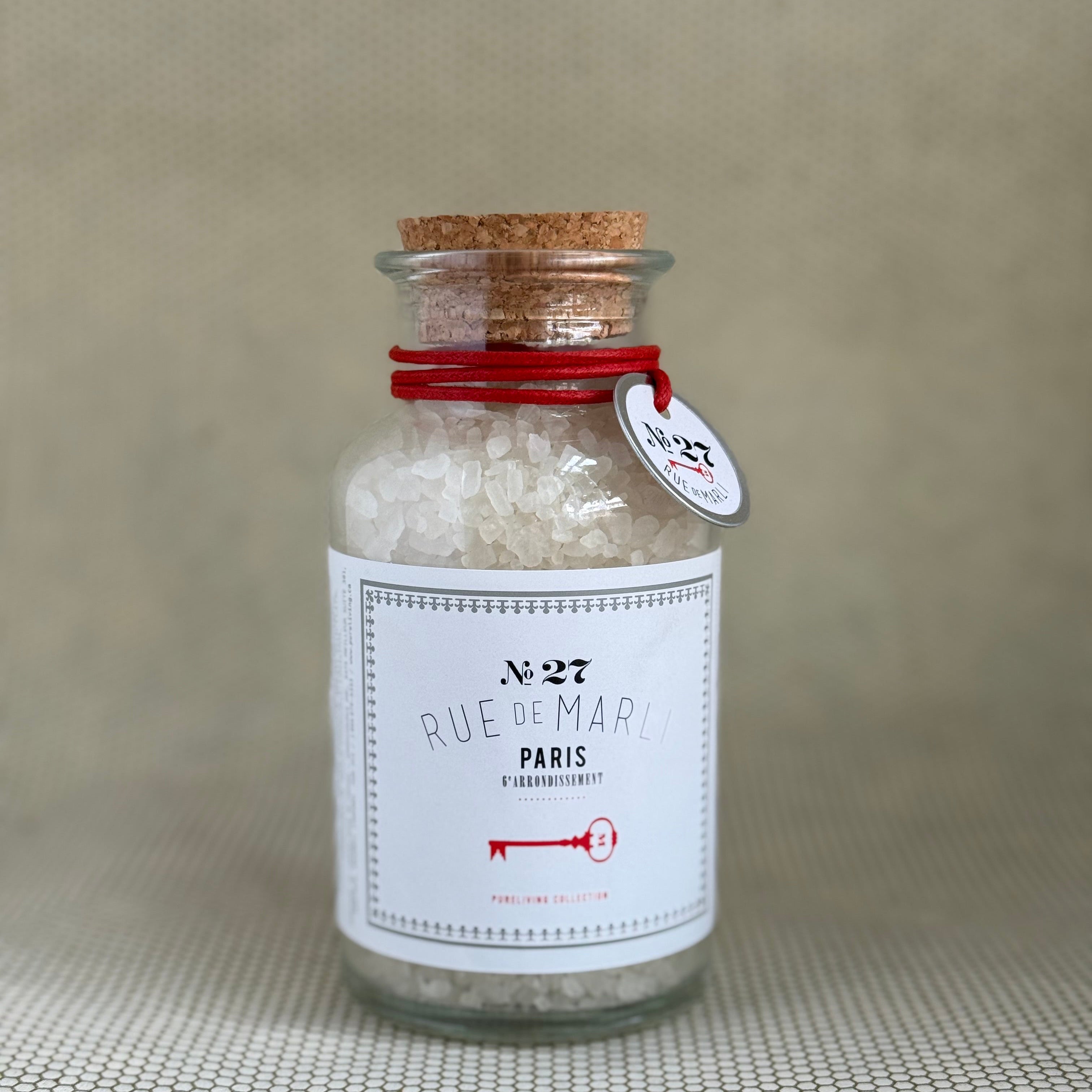 glass bottle of rue de marli bath salts with cork top and white label on neutral background. 