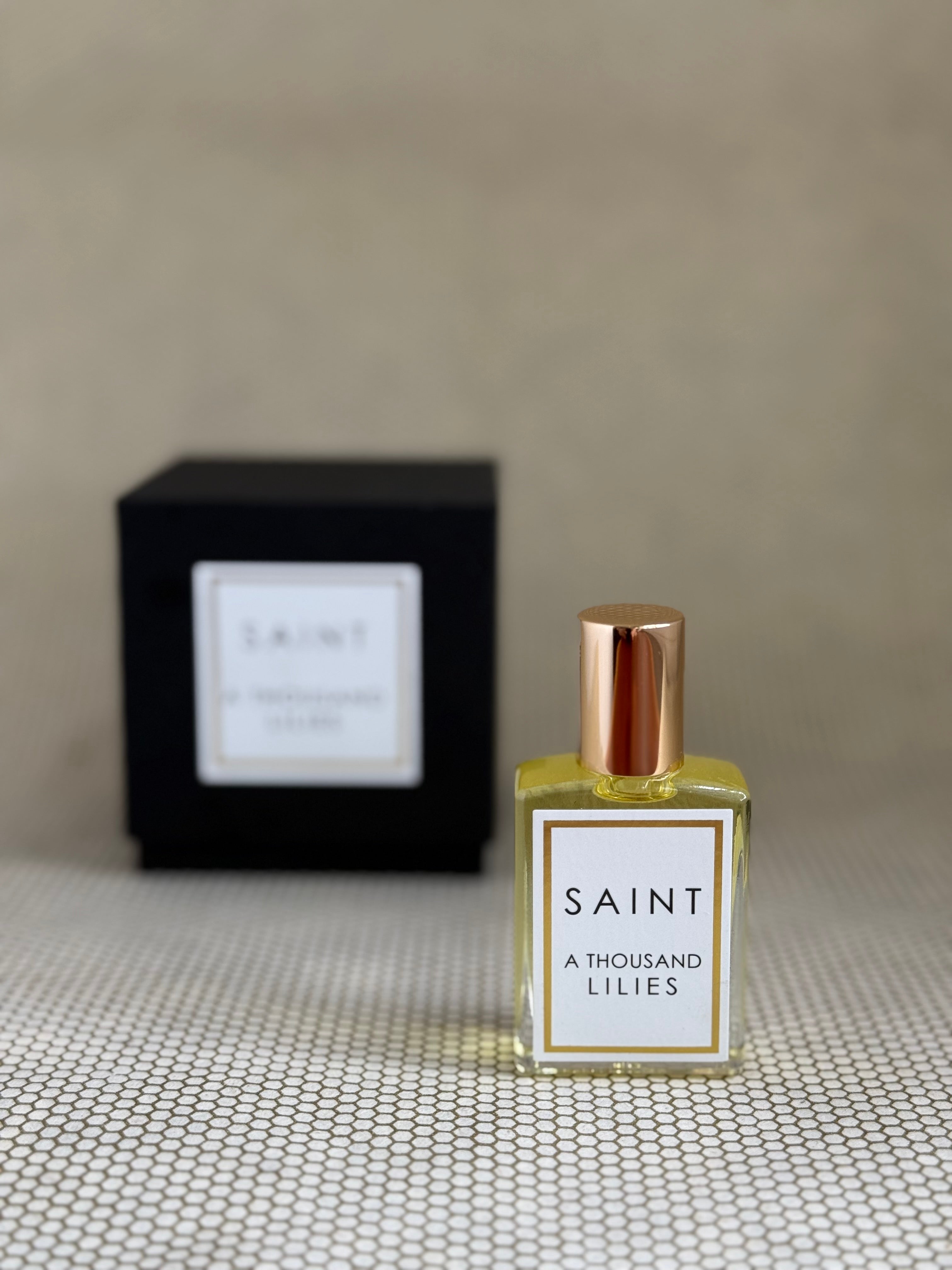 Perfume bottle labeled 'Saint A Thousand Lilies' with a black box in the background on a textured surface.