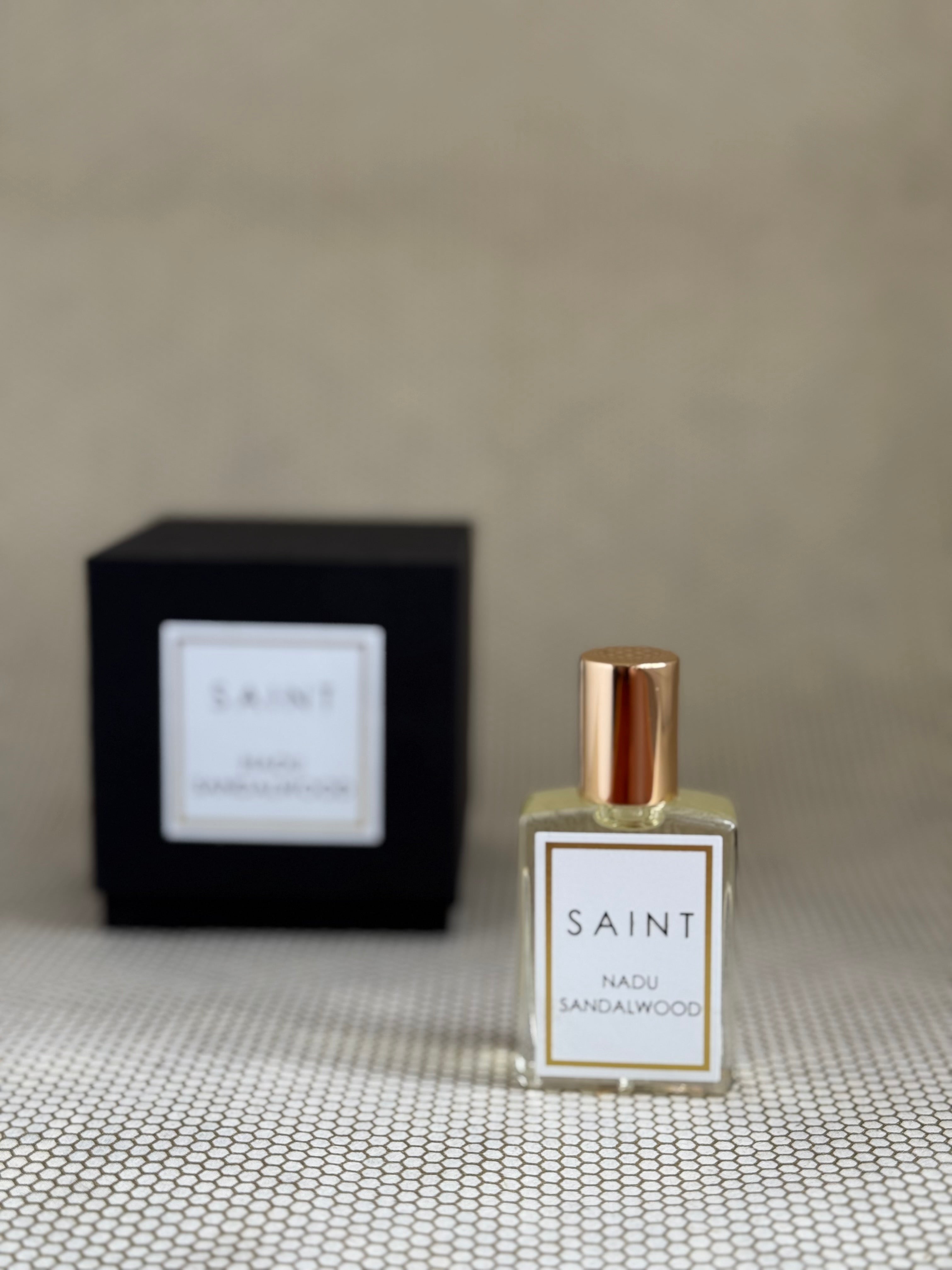 Perfume bottle labeled 'SAINT Nadu Sandalwood' with a matching black box on a textured surface.