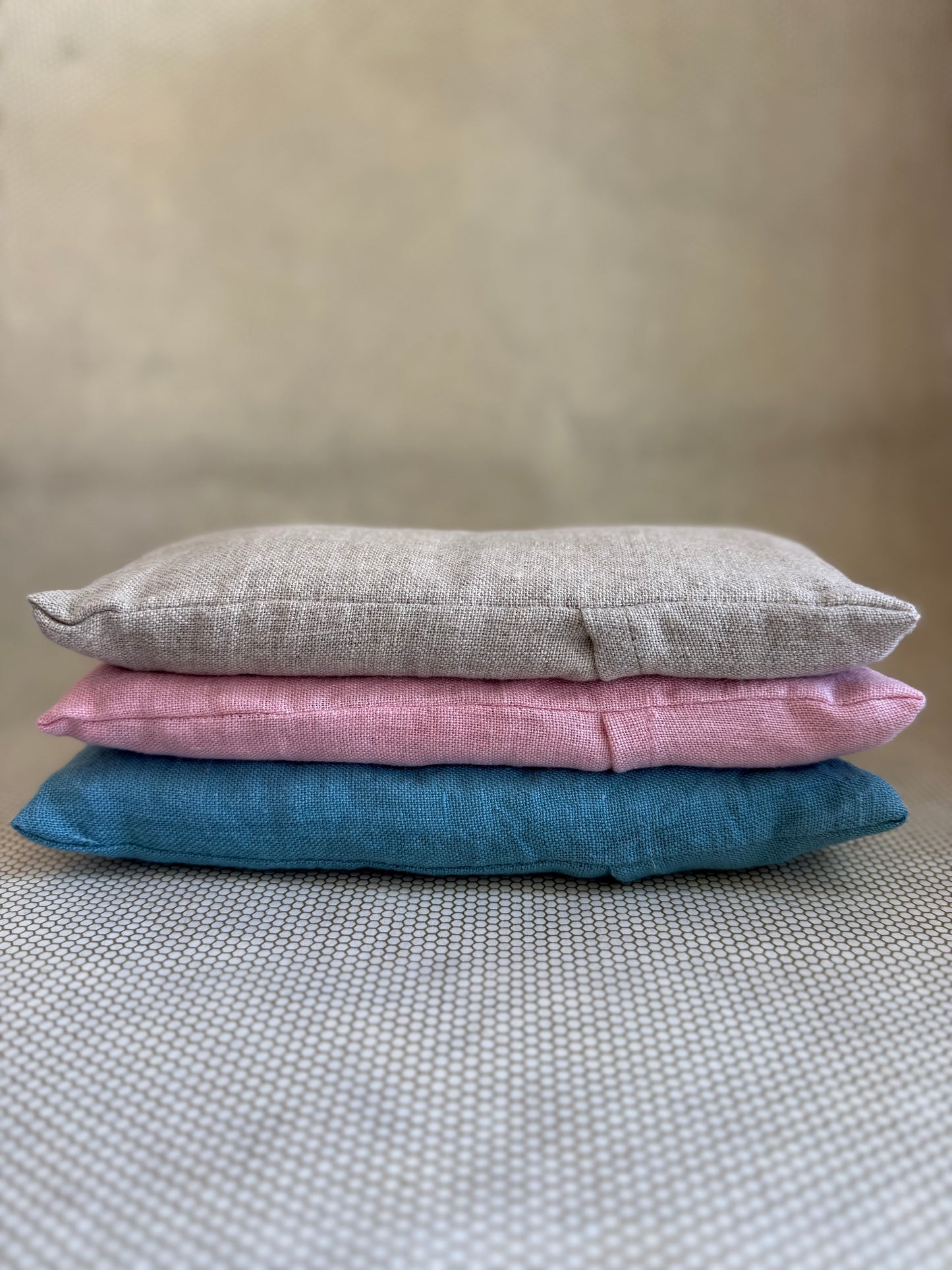 Three stacked eye pillow cushions in pink, gray, and blue on a textured surface.