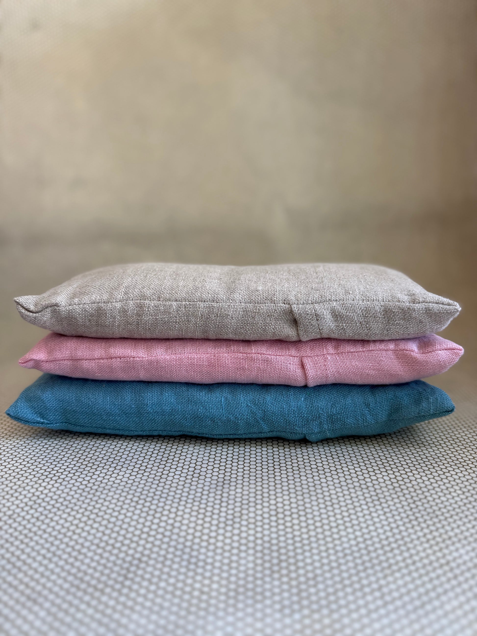 Three stacked eye pillow cushions in pink, gray, and blue on a textured surface.