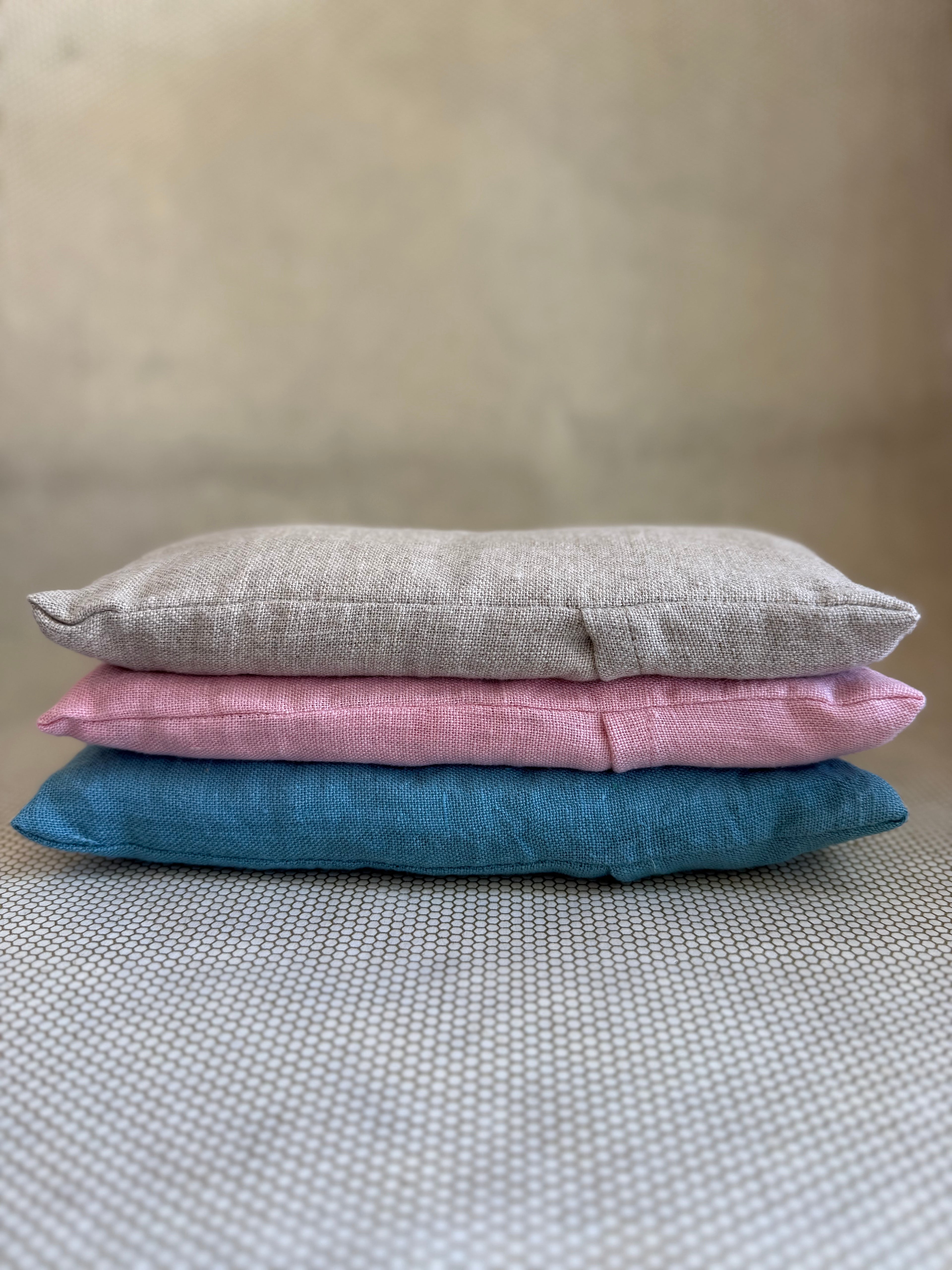 Three stacked eye pillow cushions in pink, gray, and blue on a textured surface.