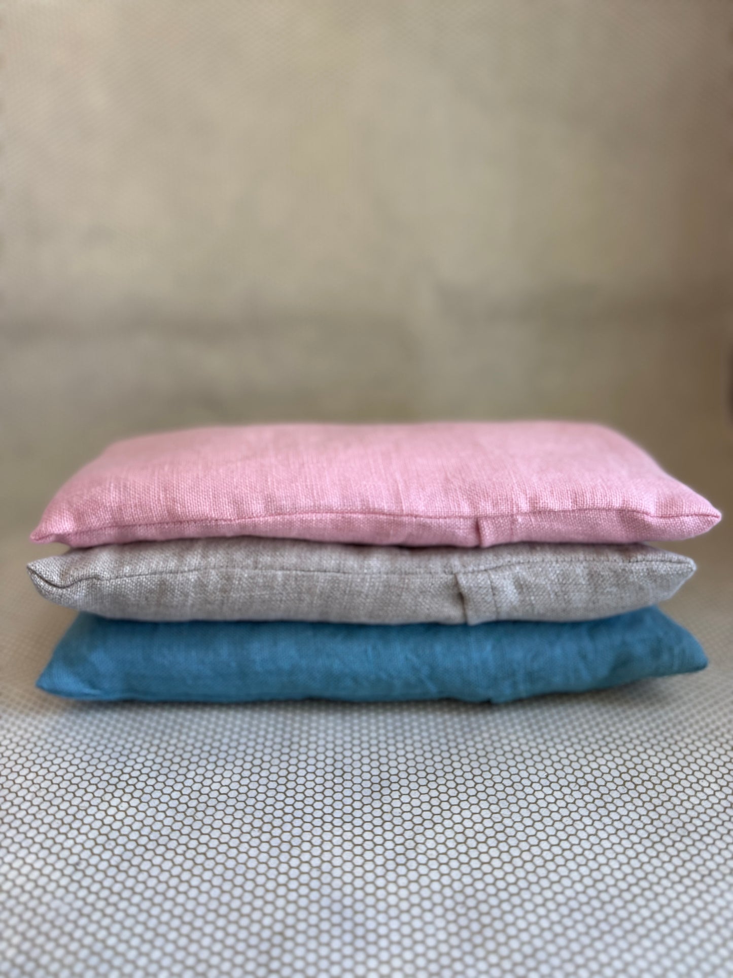 Three stacked eye pillow cushions in pink, gray, and blue on a textured surface.