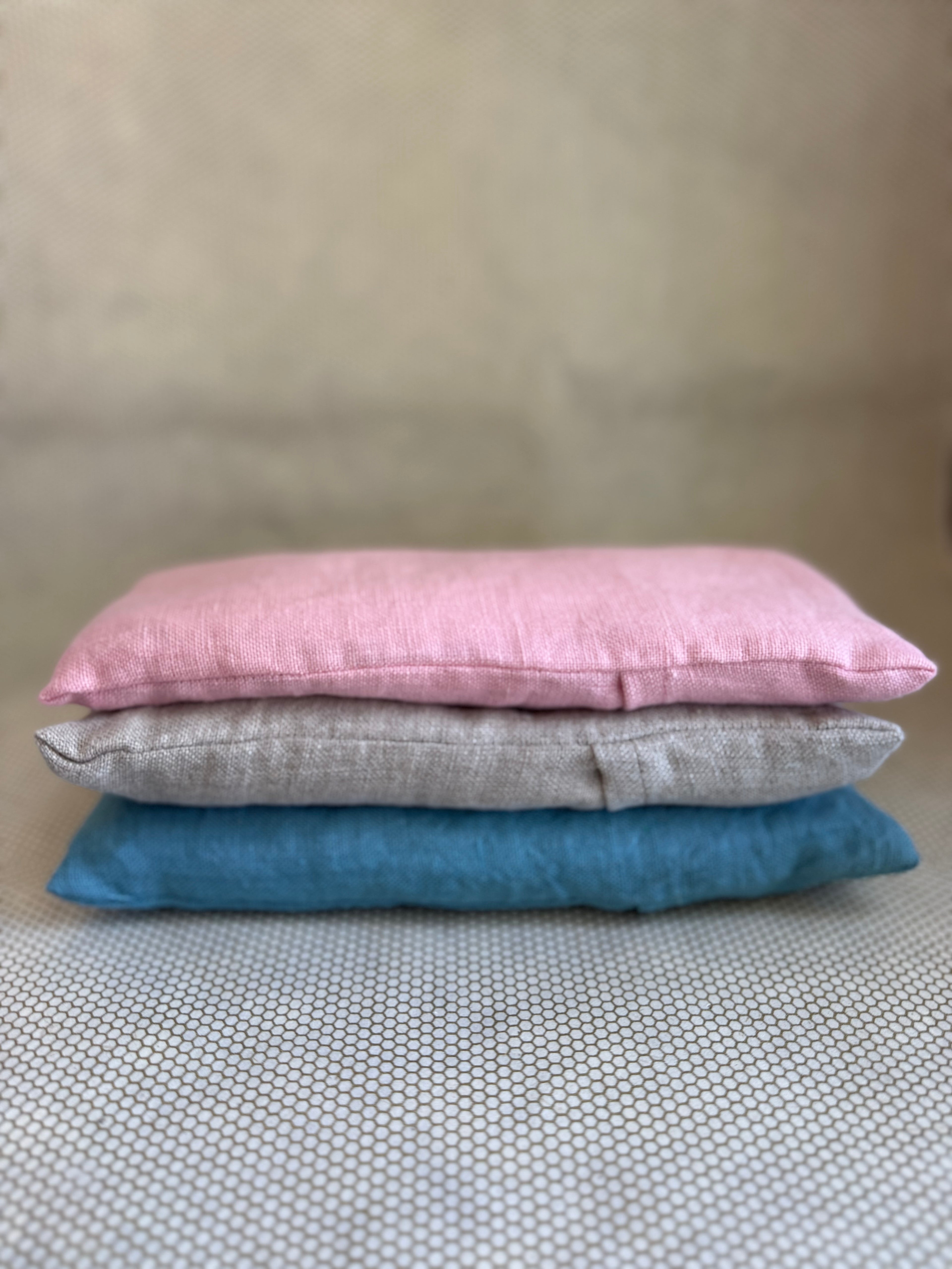 Three stacked eye pillow cushions in pink, gray, and blue on a textured surface.