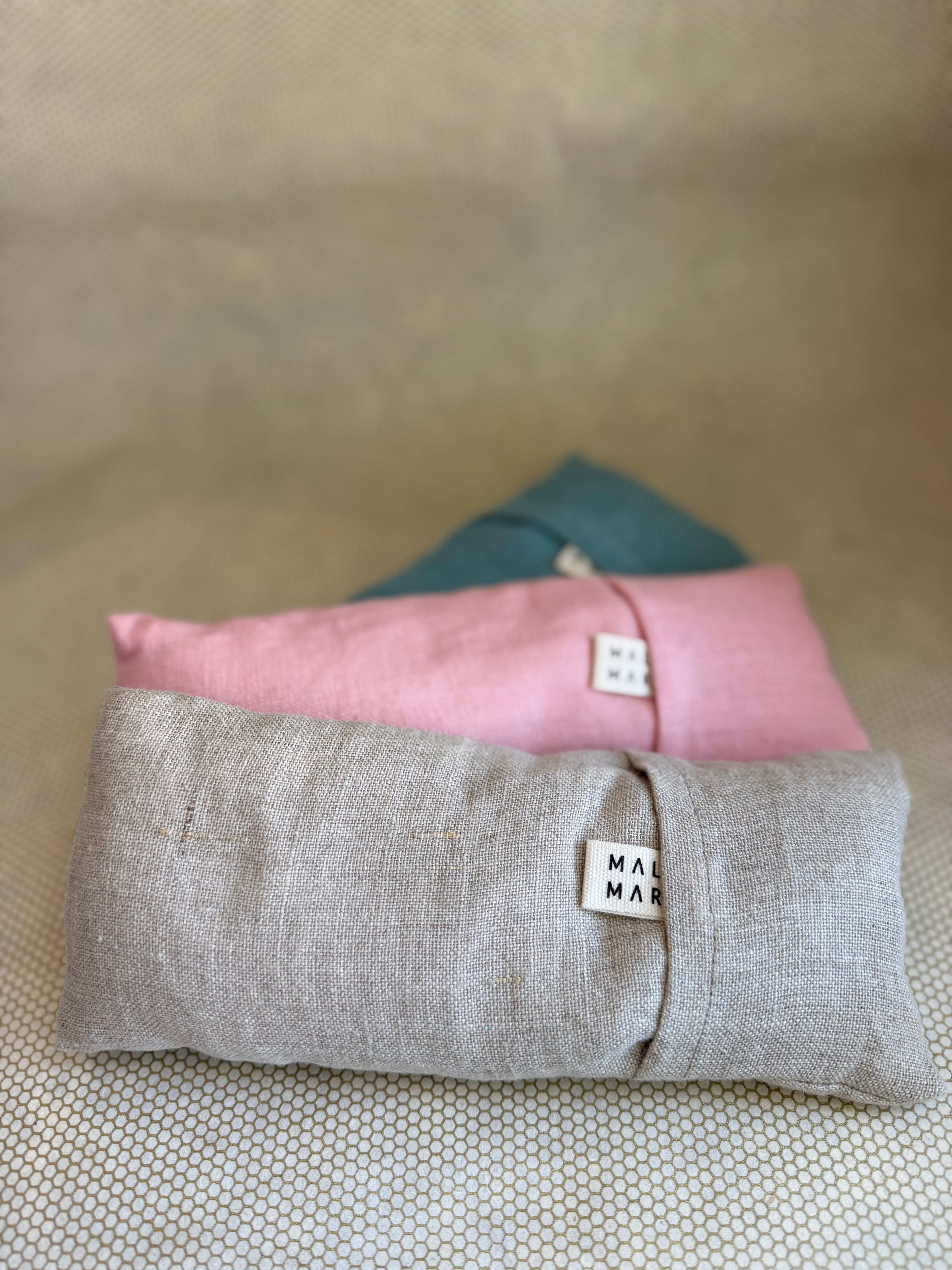 Three stacked eye pillow cushions in pink, gray, and blue on a textured surface.
