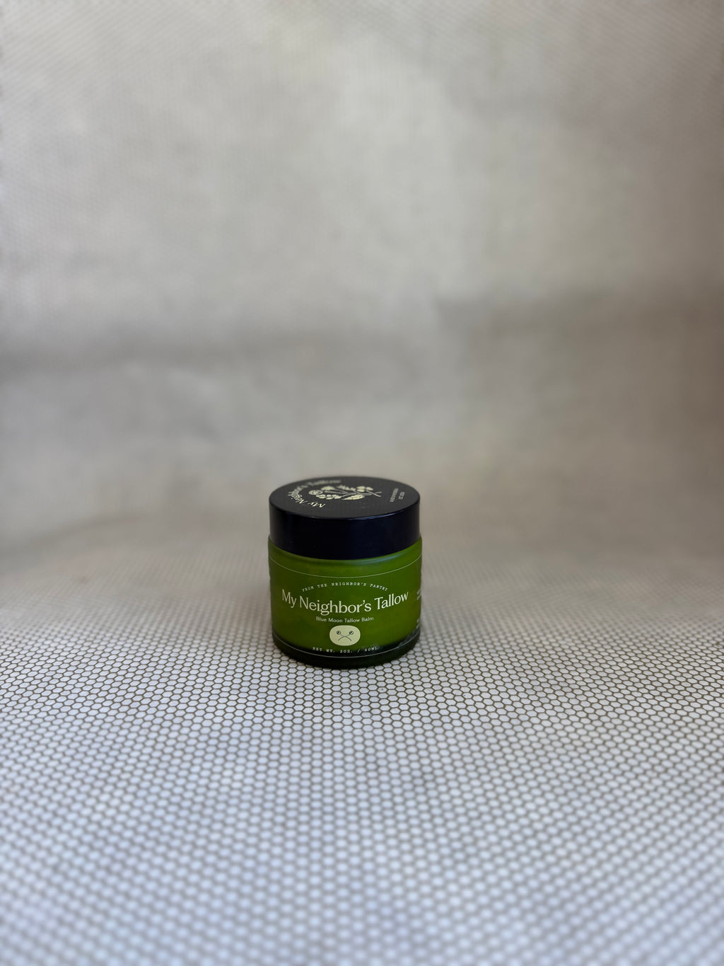 Green jar of My Neighbor's blue moon tallow balm with black twist off lid on neutral background. 