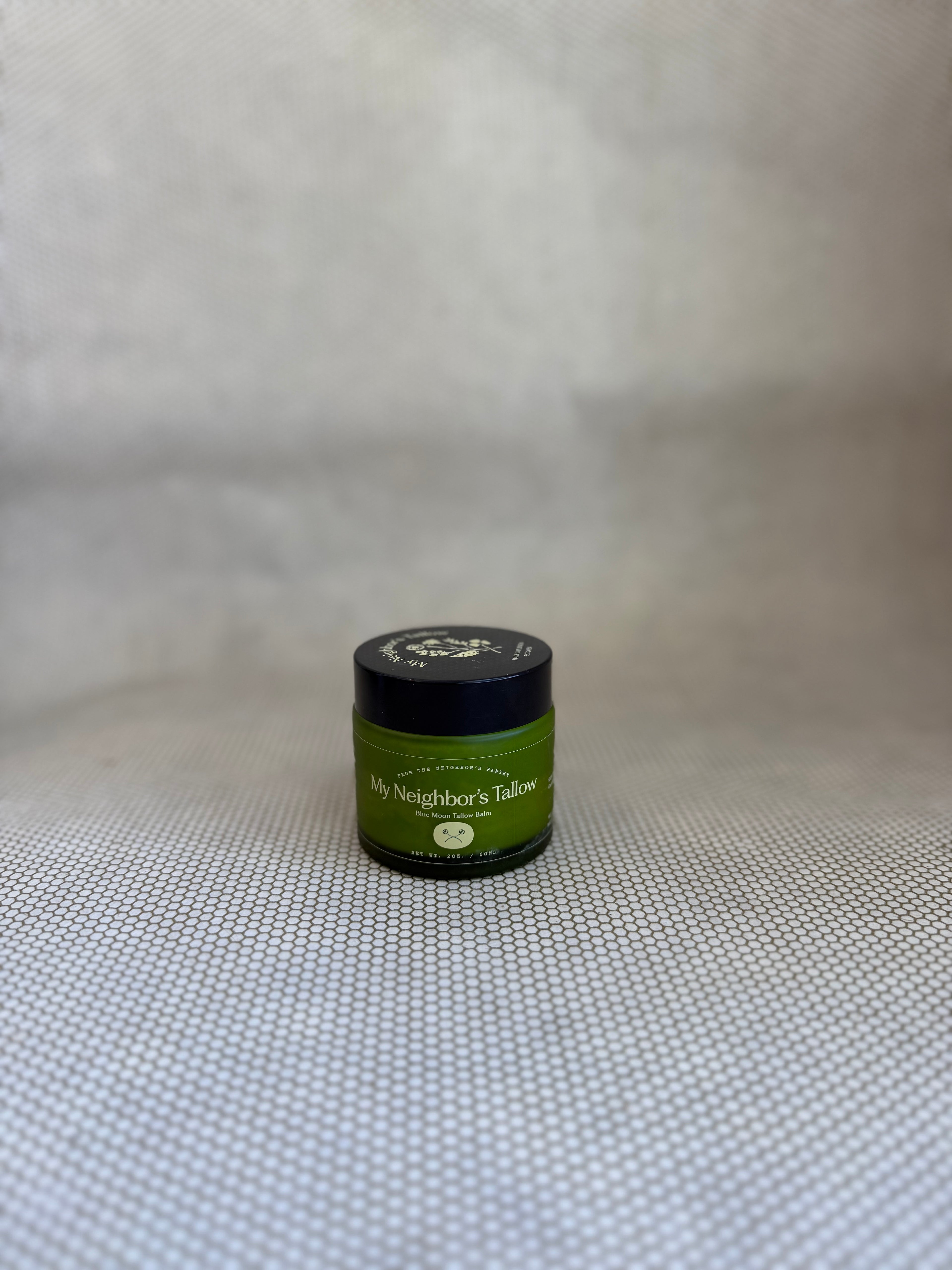Green jar of My Neighbor's blue moon tallow balm with black twist off lid on neutral background. 