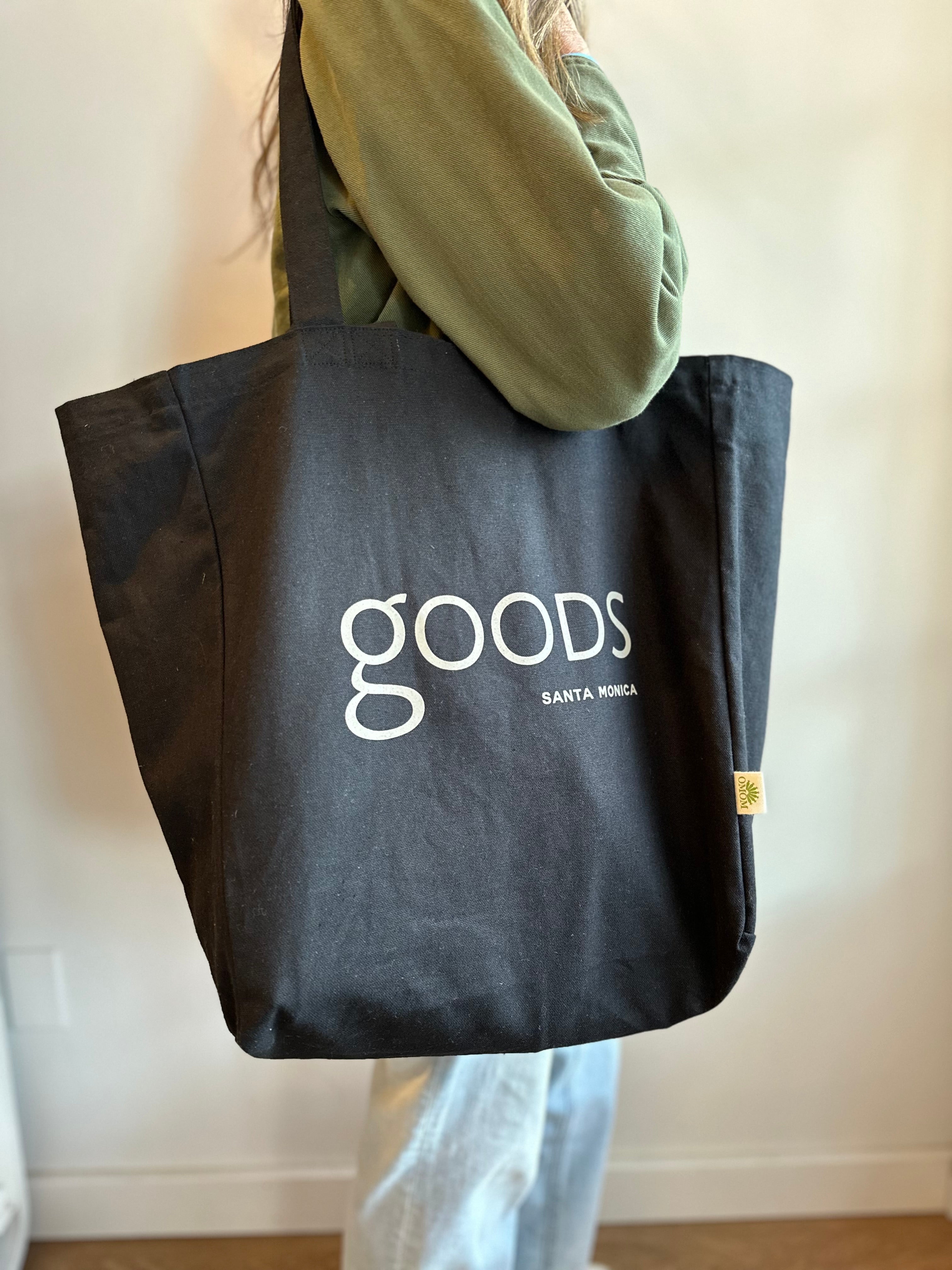 A person holding a black cotton tote bag with the brand name 'GOODS' printed in white on the front.