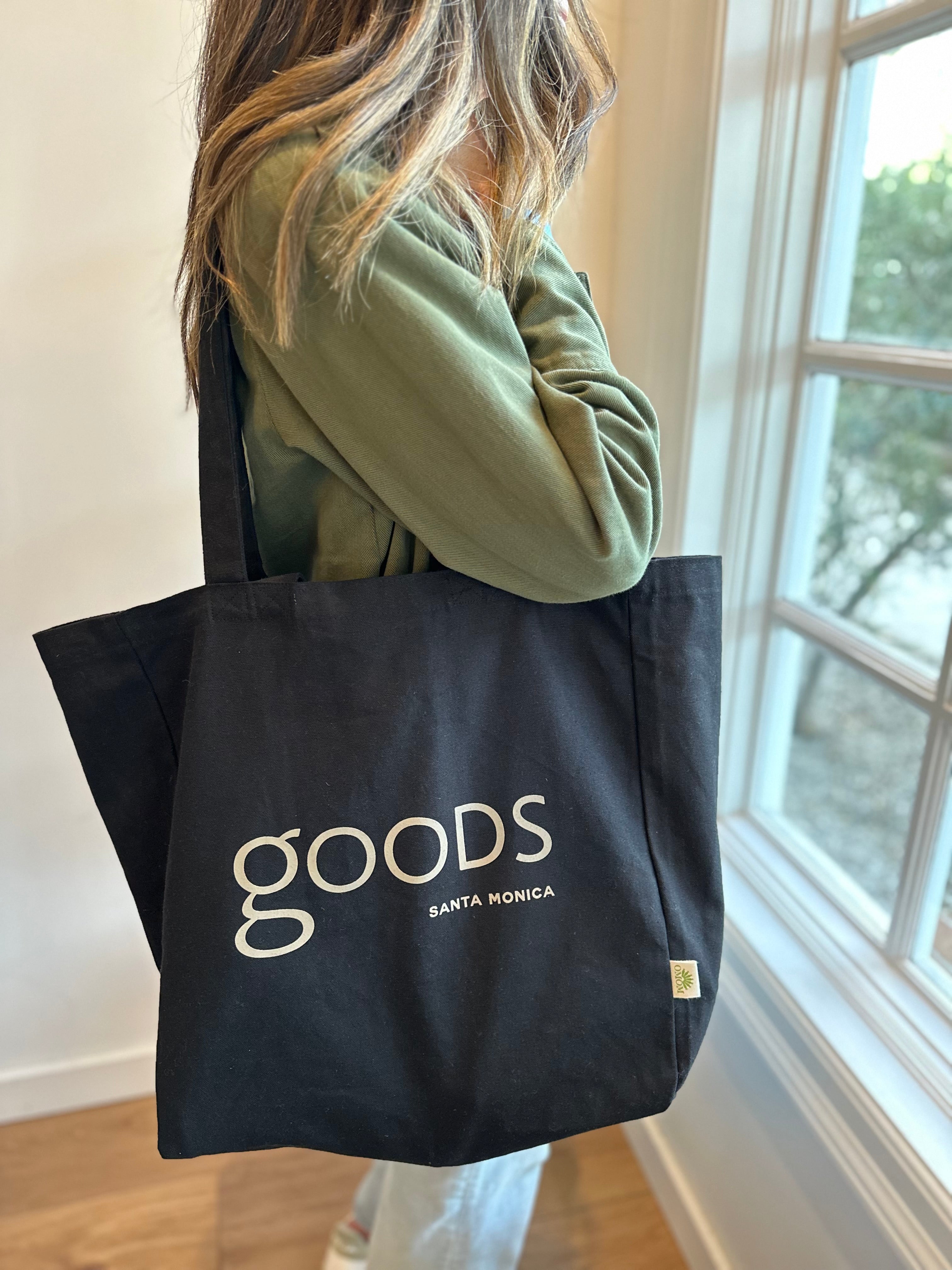 A person holding a black cotton tote bag with the brand name 'GOODS' printed in white on the front.