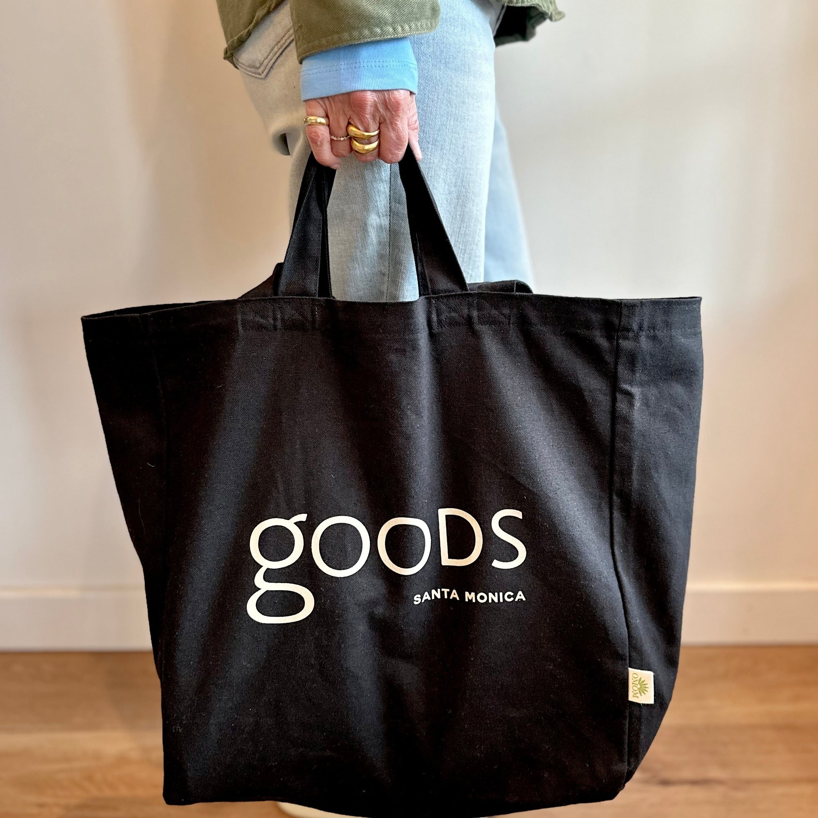 A person holding a black cotton tote bag with the brand name 'GOODS' printed in white on the front.