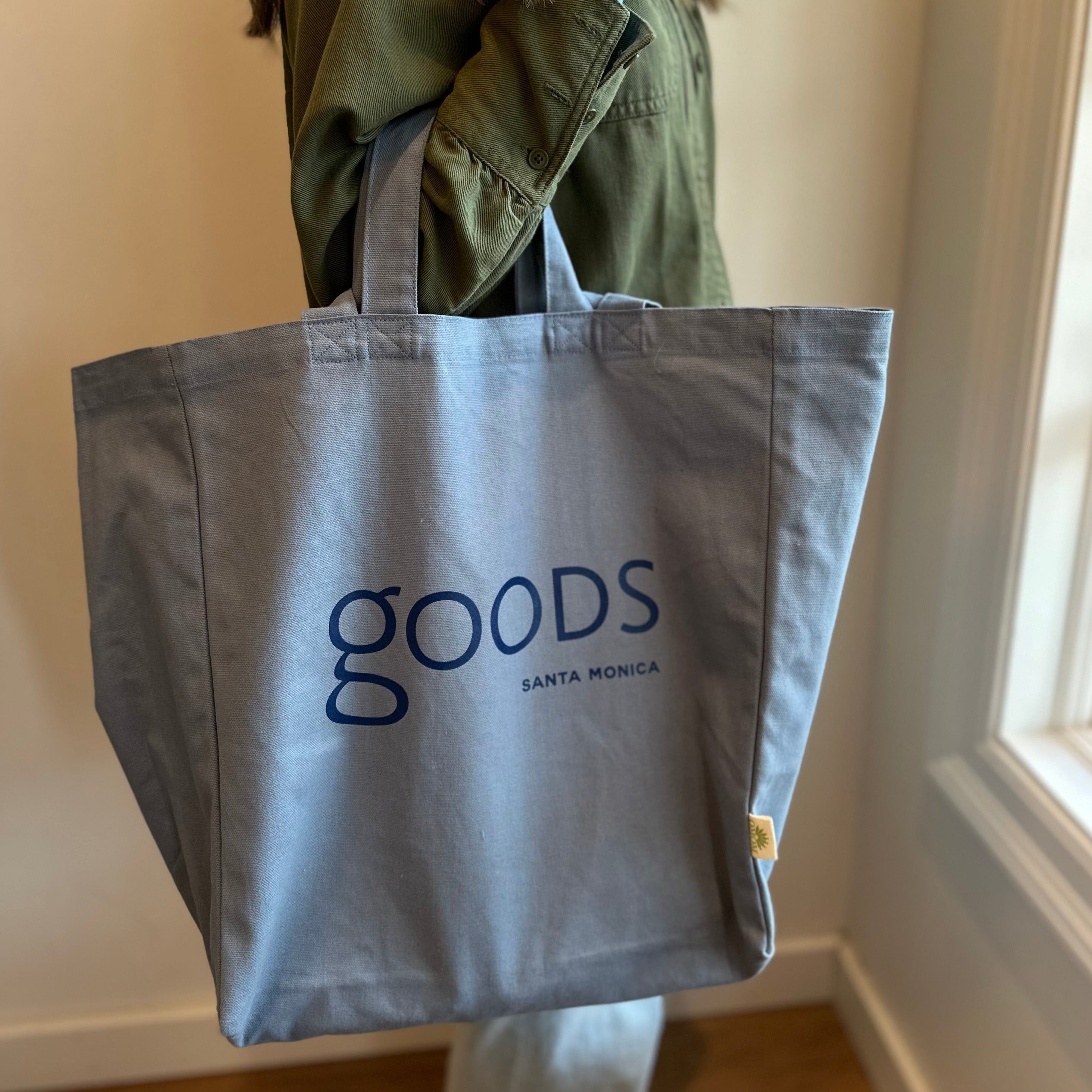 A person holding a gray cotton tote bag with the brand name 'GOODS' printed in blue letters.