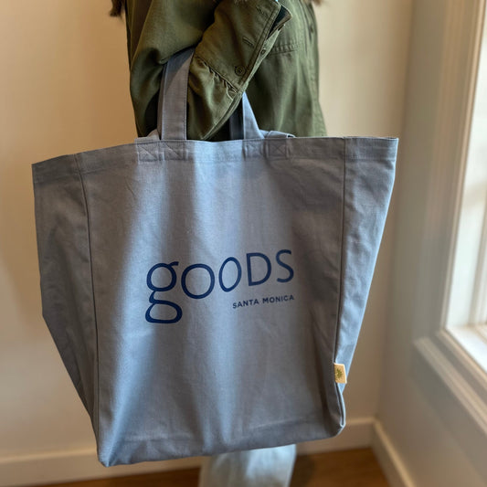 A person holding a gray cotton tote bag with the brand name 'GOODS' printed in blue letters.
