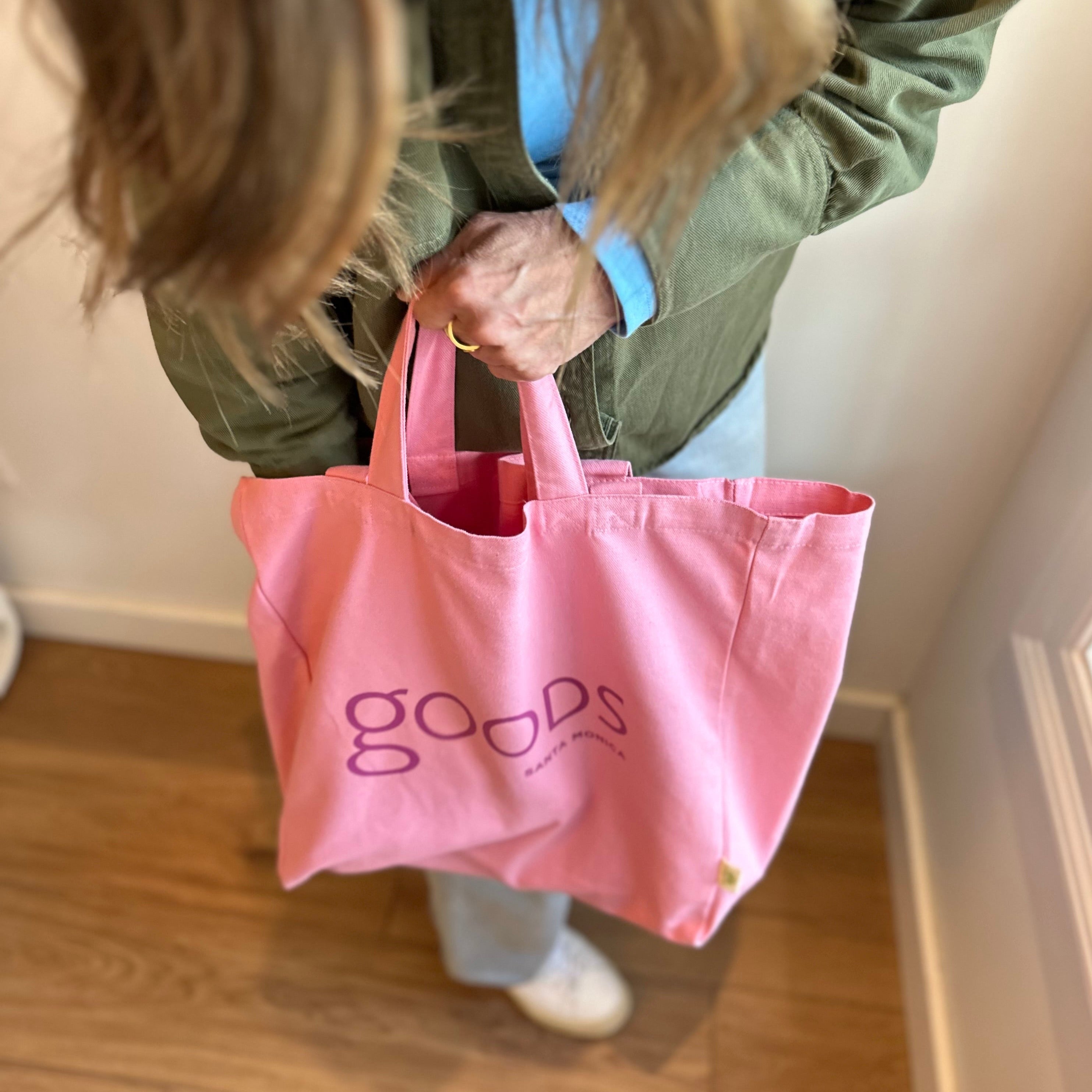 A person holding a pink cotton tote bag with the brand name 'GOODS' printed on it.