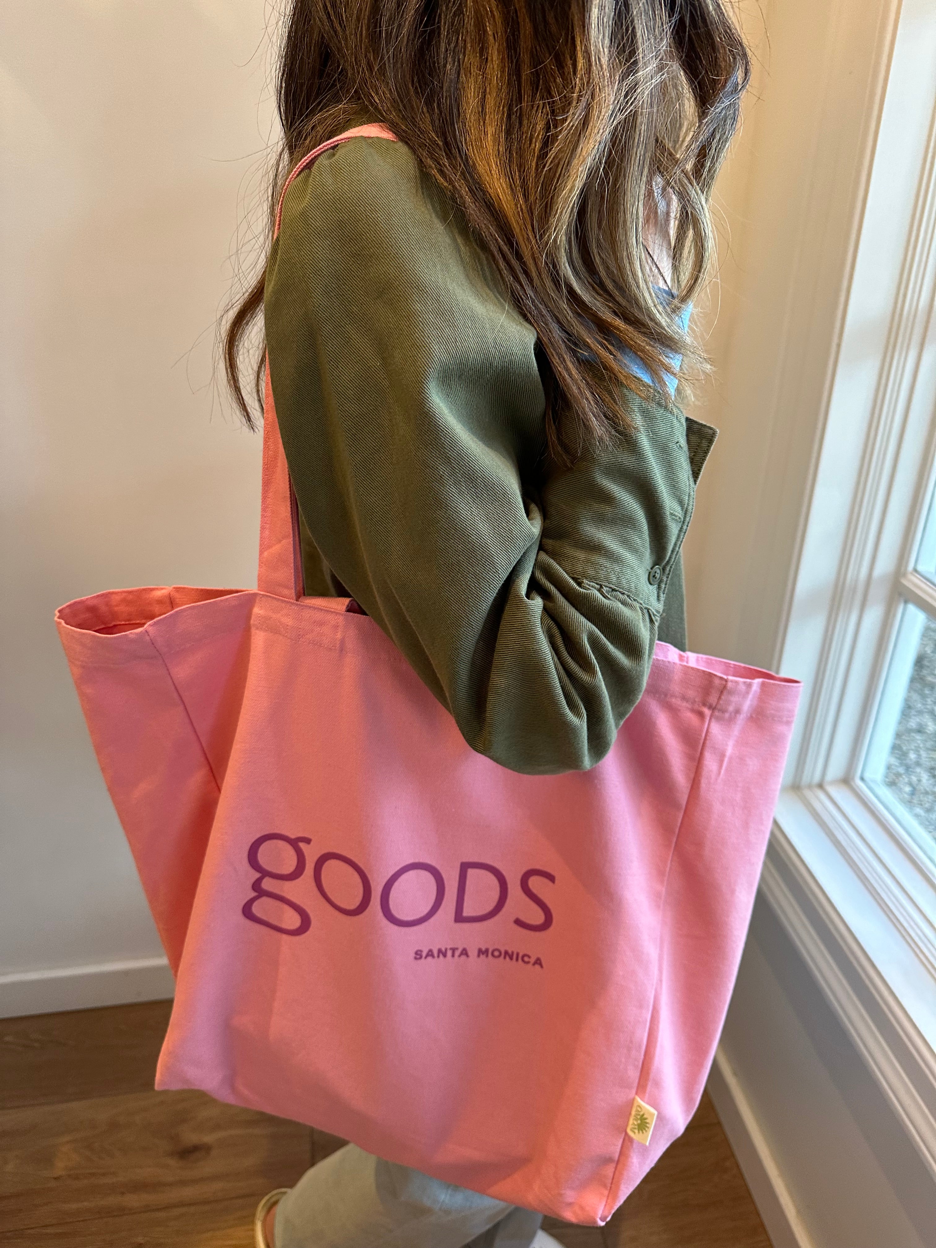 A person holding a pink cotton tote bag with the brand name 'GOODS' printed on it.
