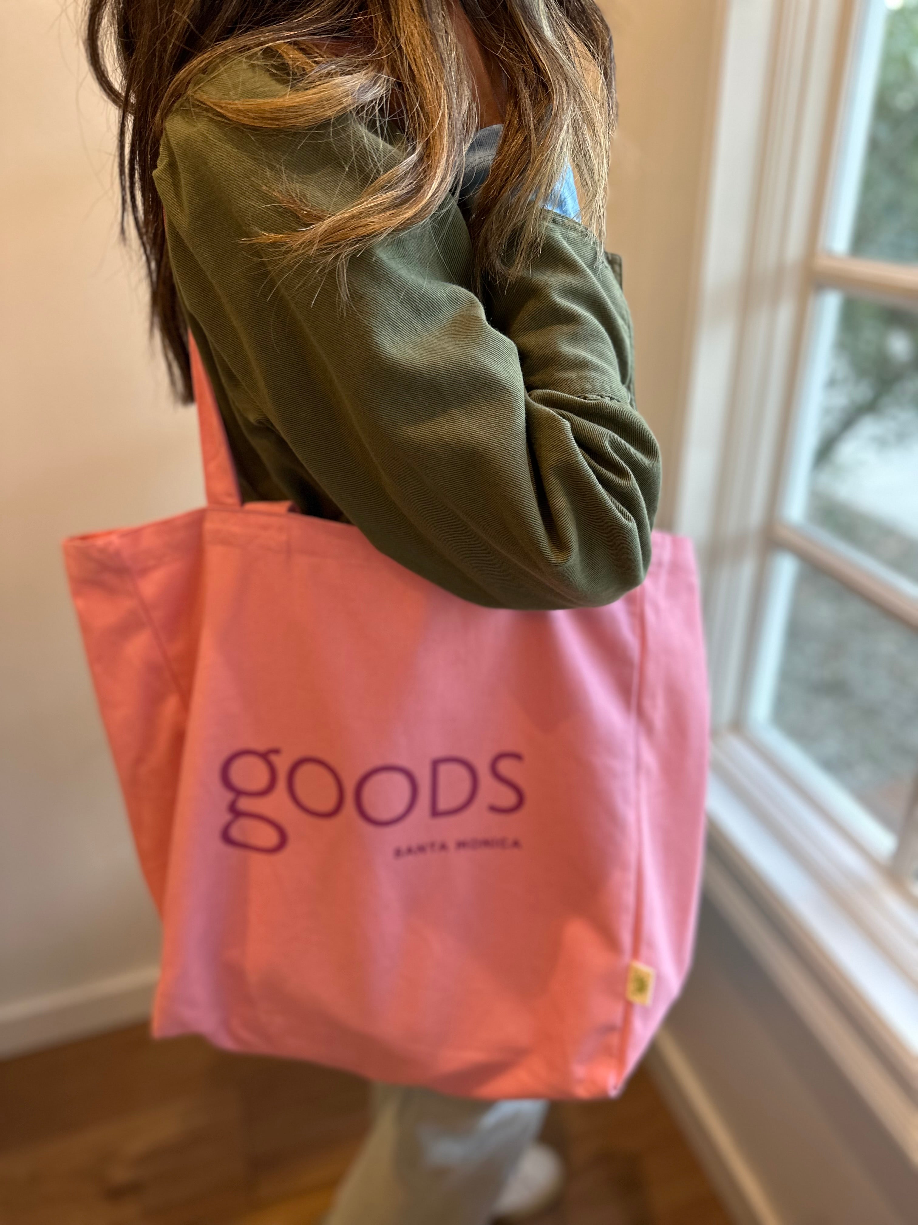 A person holding a pink cotton tote bag with the brand name 'GOODS' printed on it.
