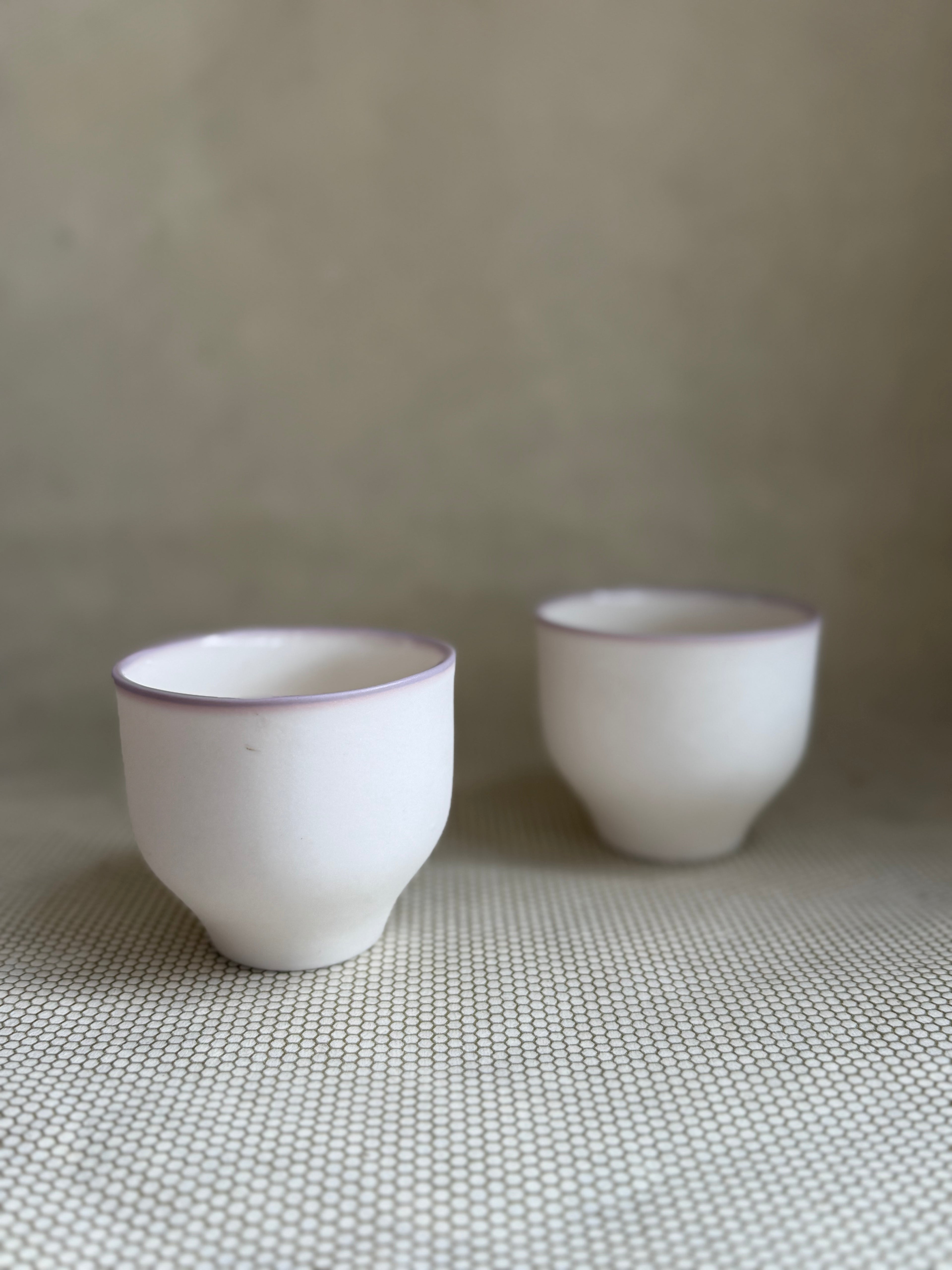 Two white porcelain cups with lavender rims on a surface with a neutral background.